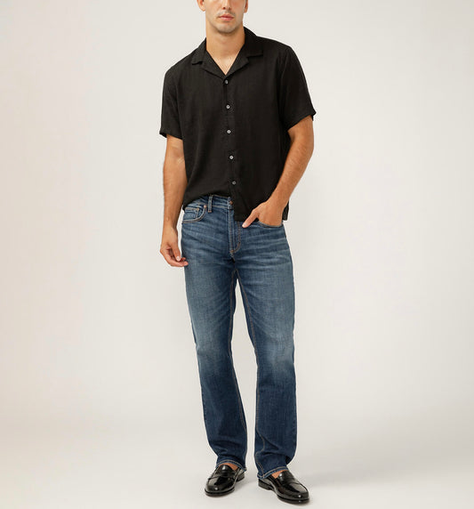 A man wearing a black short-sleeved button-up shirt and Silver Jeans Co. Grayson Classic Fit Straight Leg Jeans in Indigo stands with one hand in his pocket. His black shoes complement the outfit against the plain light gray background.