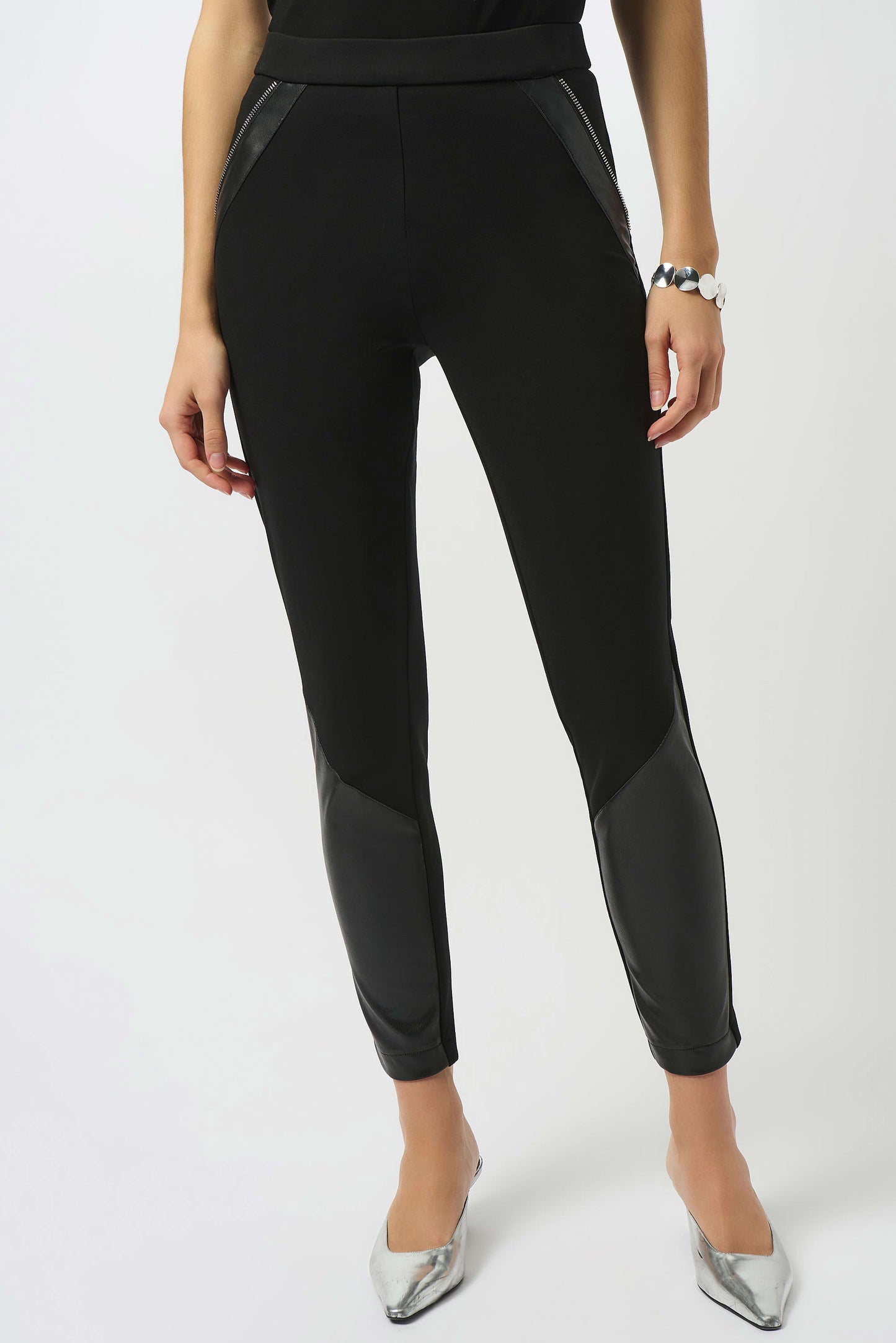 A woman wears Joseph Ribkoff's Heavy Knit and Leatherette Pull-On Legging 253078 with leather-like thigh and calf panels, paired with a bracelet and metallic silver pointed-toe heels, standing against a plain white background.