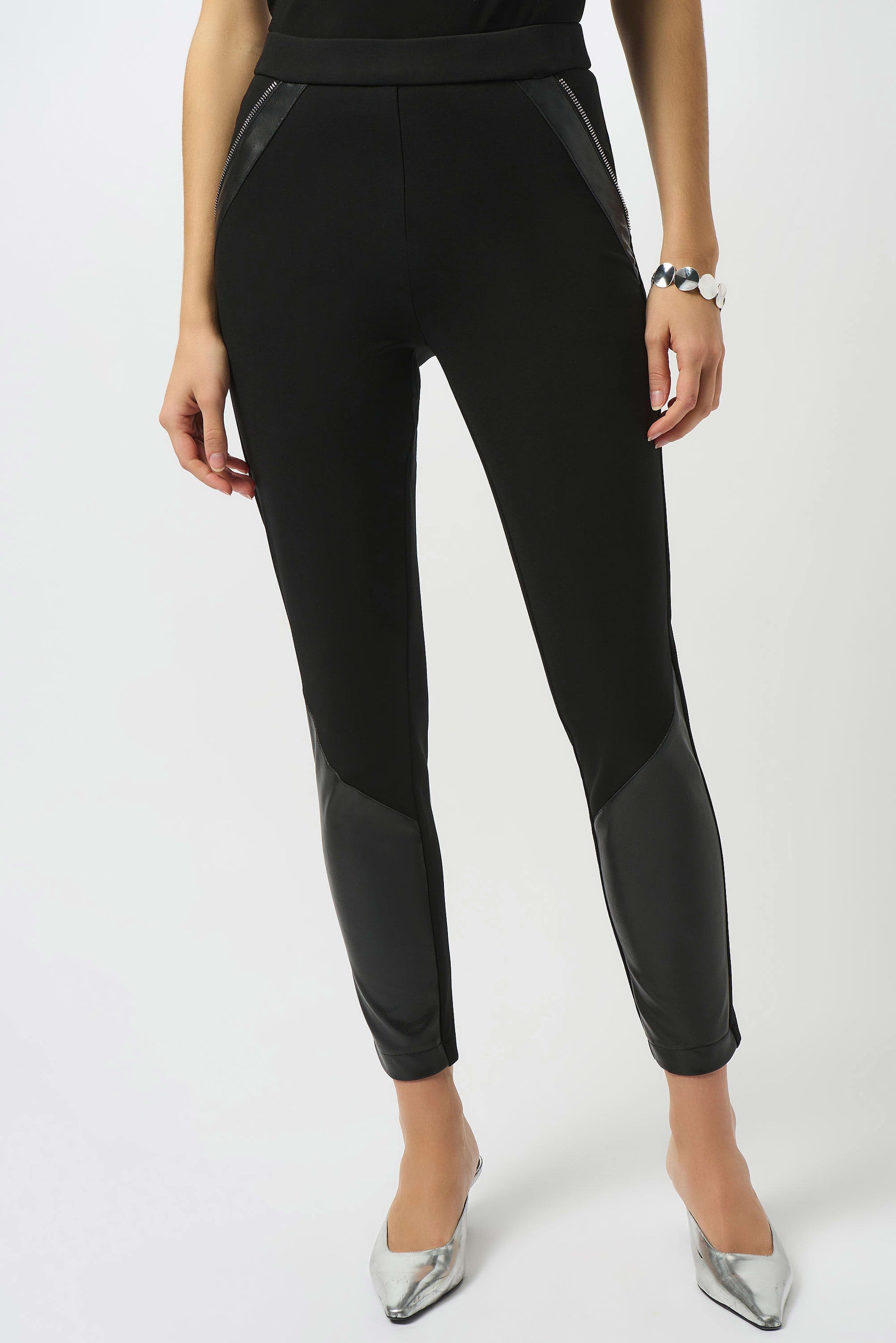 A woman wears Joseph Ribkoff's Heavy Knit and Leatherette Pull-On Legging 253078 with leather-like thigh and calf panels, paired with a bracelet and metallic silver pointed-toe heels, standing against a plain white background.