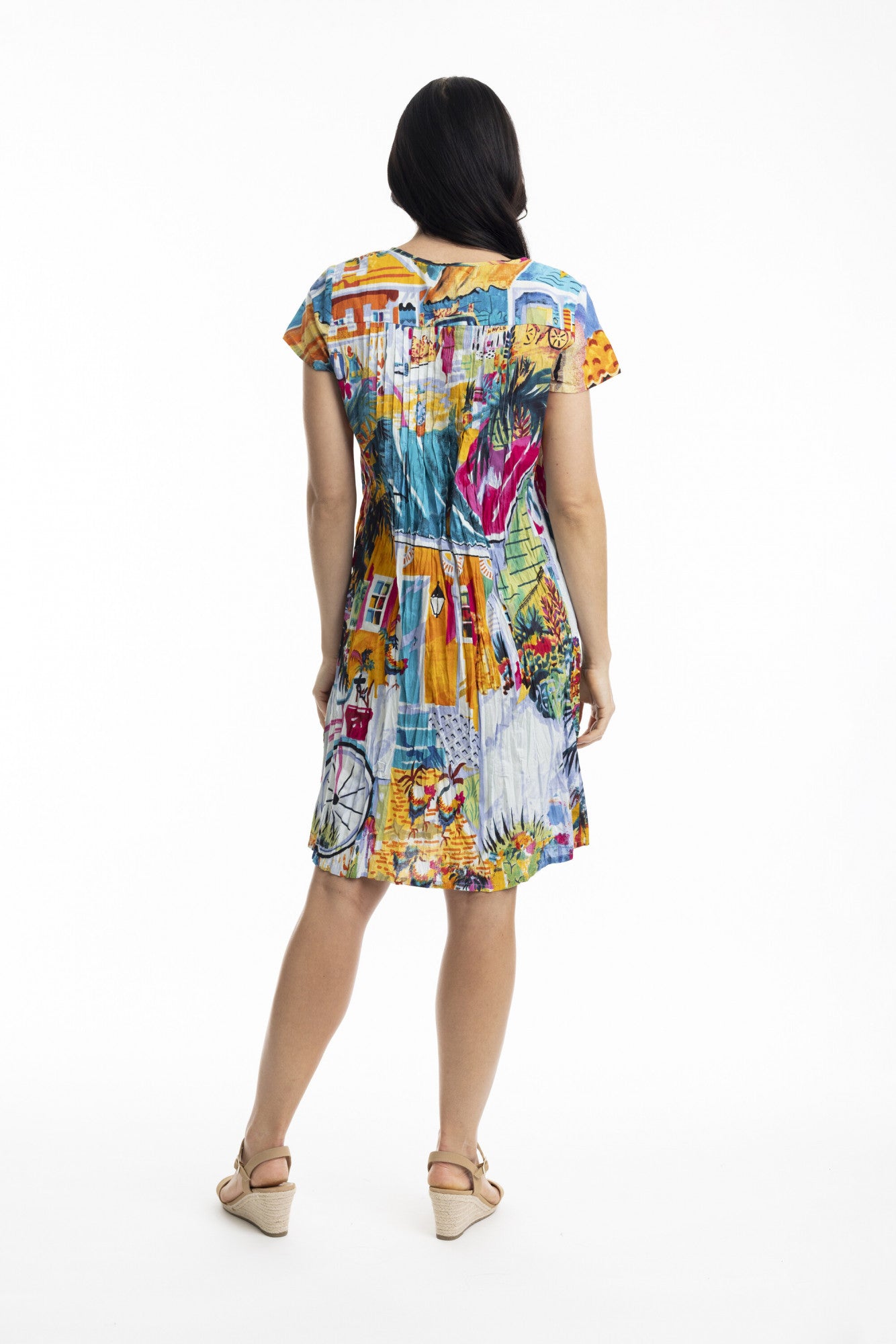 A woman with long dark hair faces away, wearing the ORIENTIQUE Printed Cotton Easy Fit Dress Style: 20058 Florida, featuring a colorful artistic print and V-neckline, paired with beige wedge sandals against a plain white background.