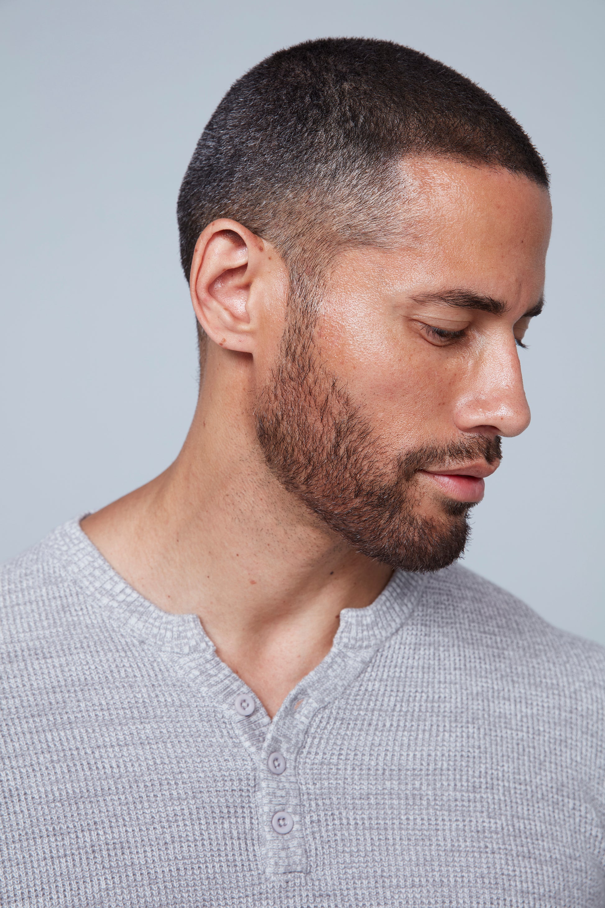 A stylish man with short hair and a beard stands against a grey background, looking down to his right with his arms crossed over his chest. He is wearing the light grey Henley Sweater Cotton from HEDGE, made from 100% cotton knit, paired with black pants.