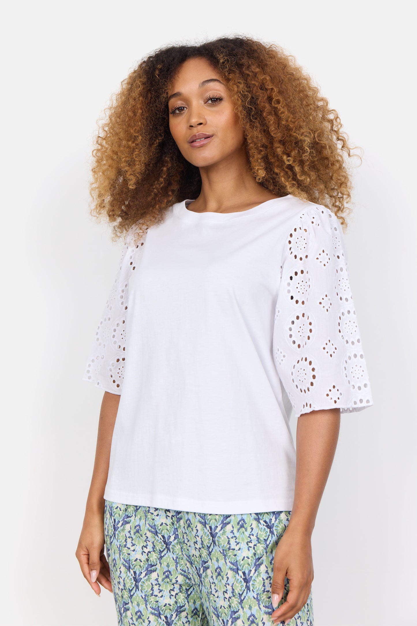 A curly-haired woman wears the stylish Loraine 3 Shirt by SoyaConcept, crafted from organic cotton, paired with patterned pants featuring blue and green floral designs. Neutral background.