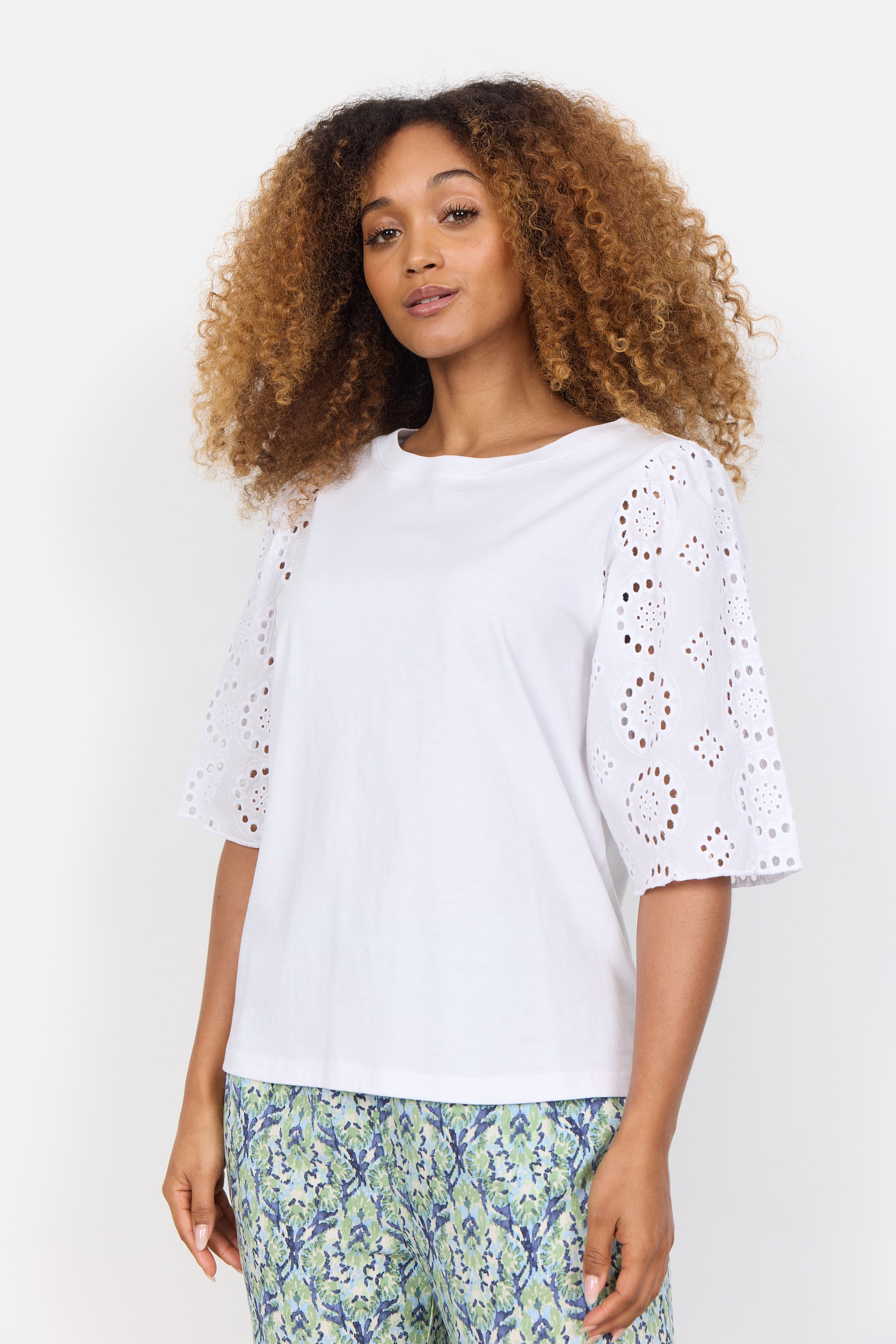 A curly-haired woman wears the stylish Loraine 3 Shirt by SoyaConcept, crafted from organic cotton, paired with patterned pants featuring blue and green floral designs. Neutral background.