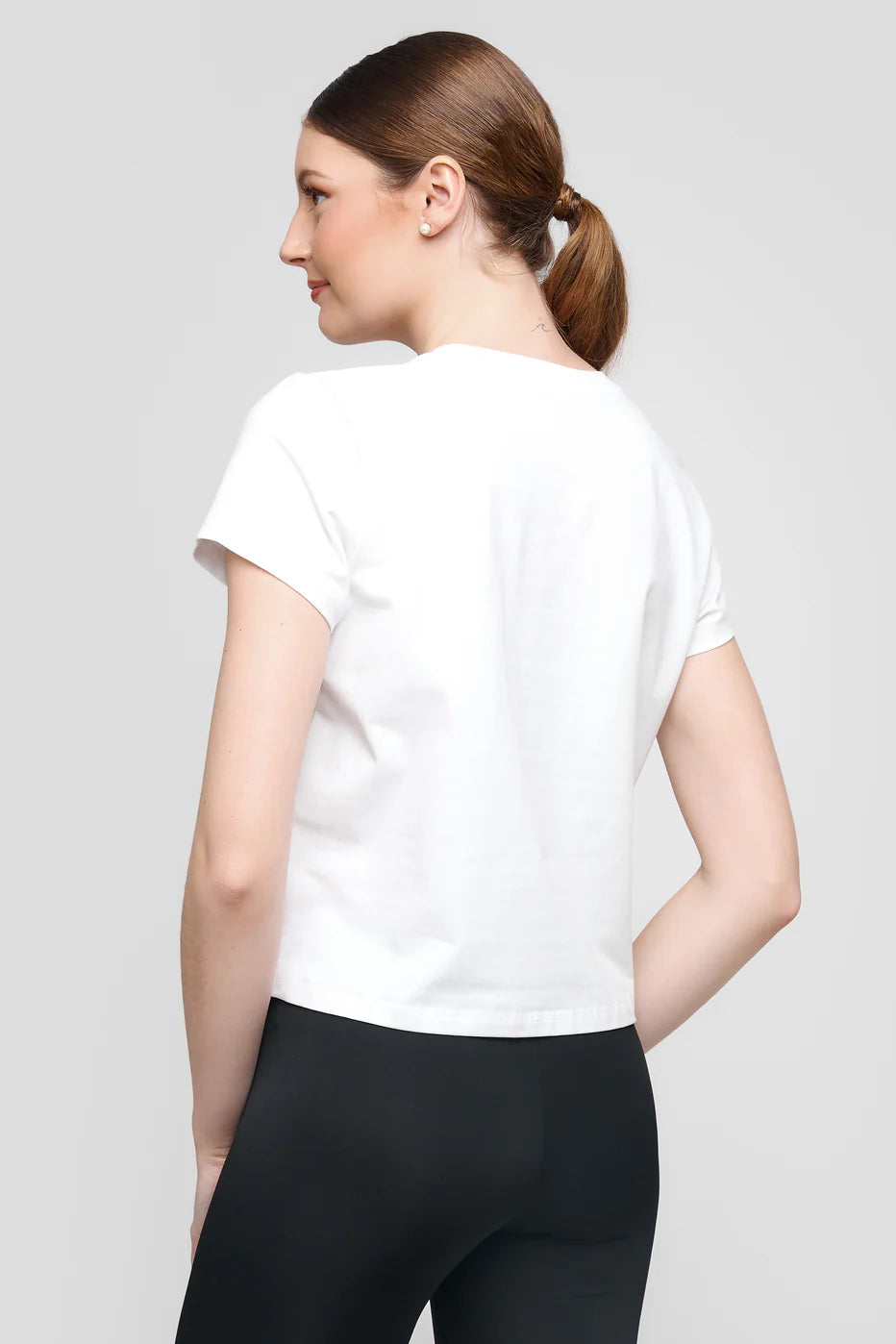 A woman with brown hair in a ponytail is wearing the UCHUU white cotton spandex t-shirt (Model: TOP CS25-303 UCHUU) and black pants, facing slightly to the side against a plain gray background.