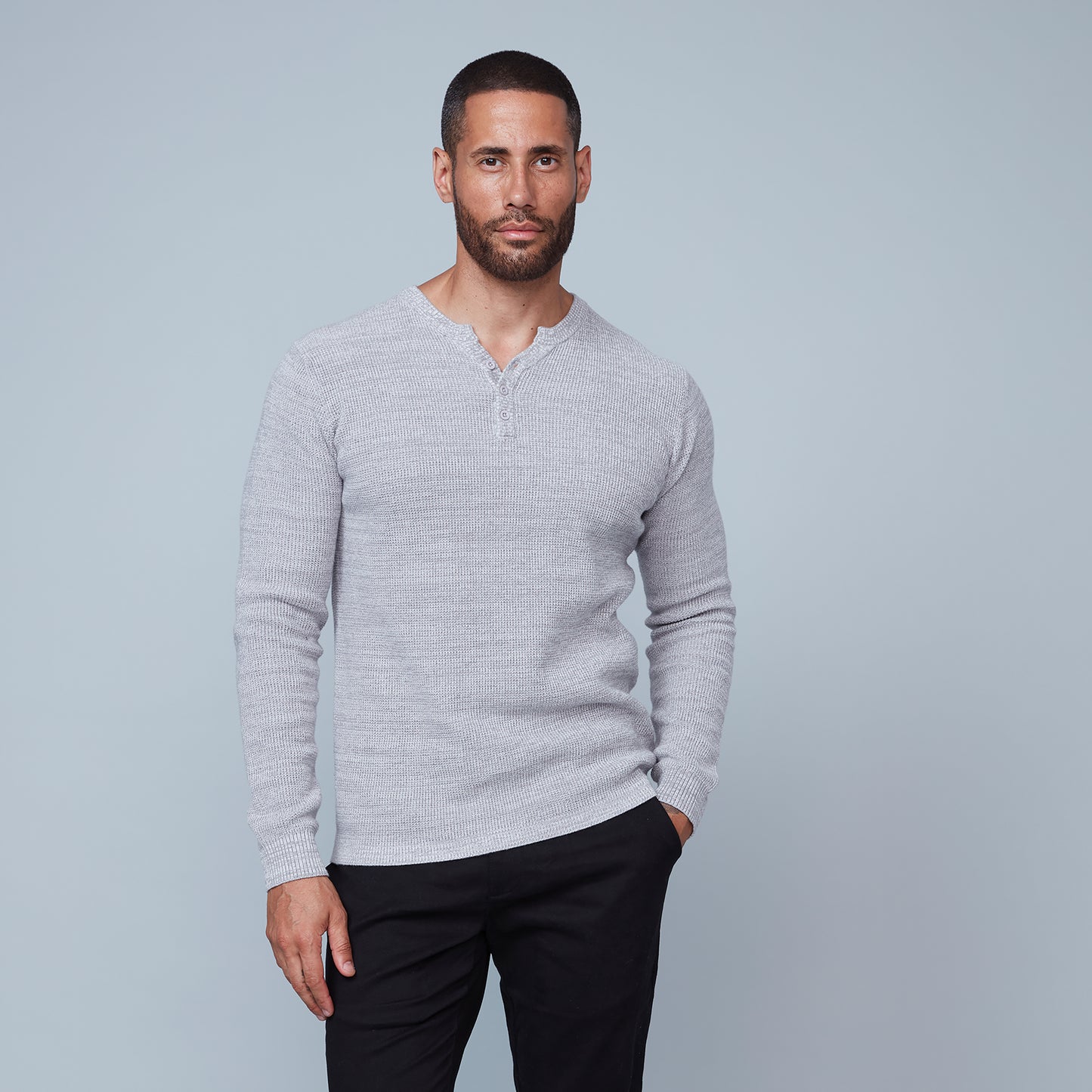 A stylish man with short hair and a beard stands against a grey background, looking down to his right with his arms crossed over his chest. He is wearing the light grey Henley Sweater Cotton from HEDGE, made from 100% cotton knit, paired with black pants.