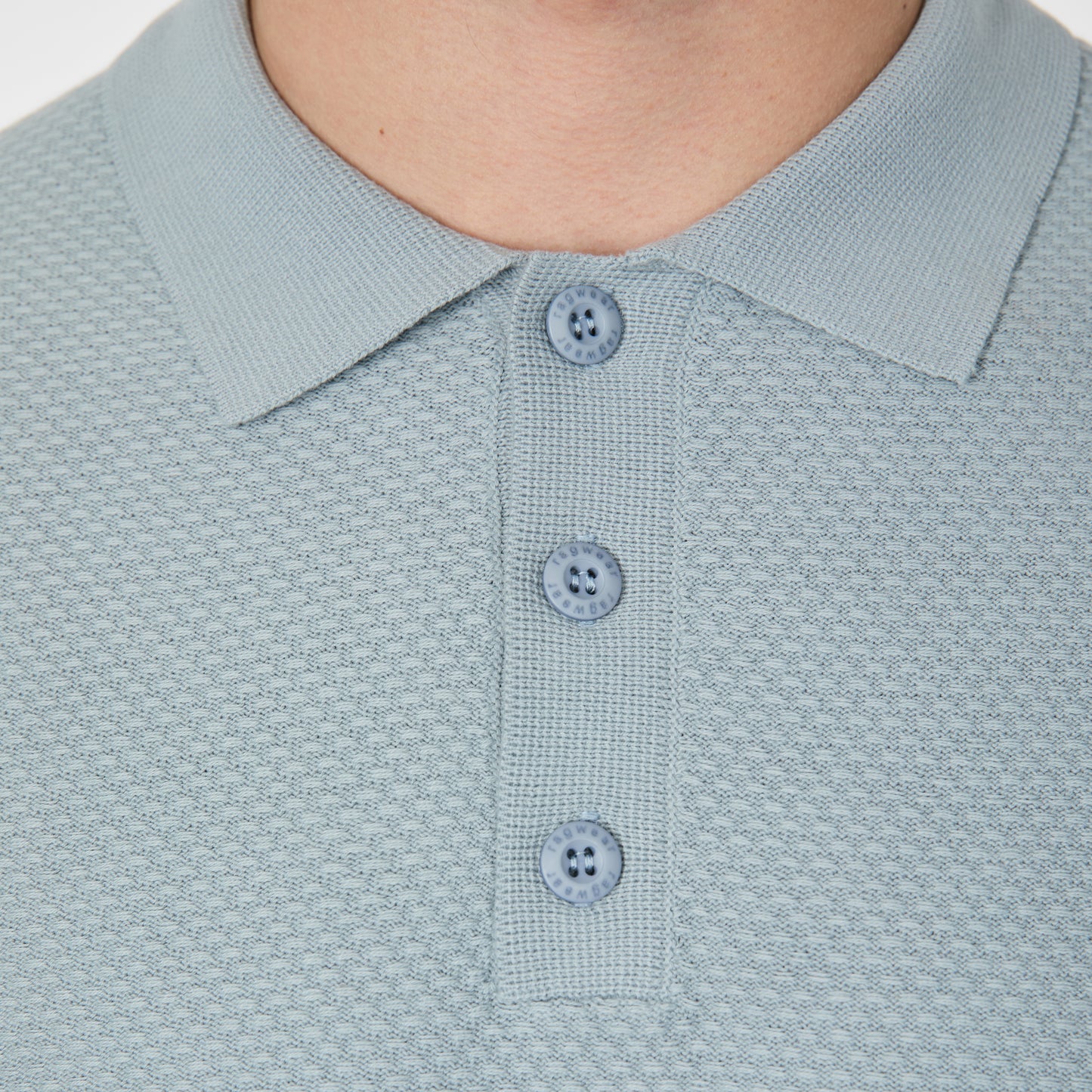 Close-up of a person wearing the RAGWEAR Polo Tarsh Short 2612-15050-2046 in Dusty Blue, featuring textured light blue 100% cotton fabric, three matching buttons, and a classic collar. Only the neck and upper chest are shown.