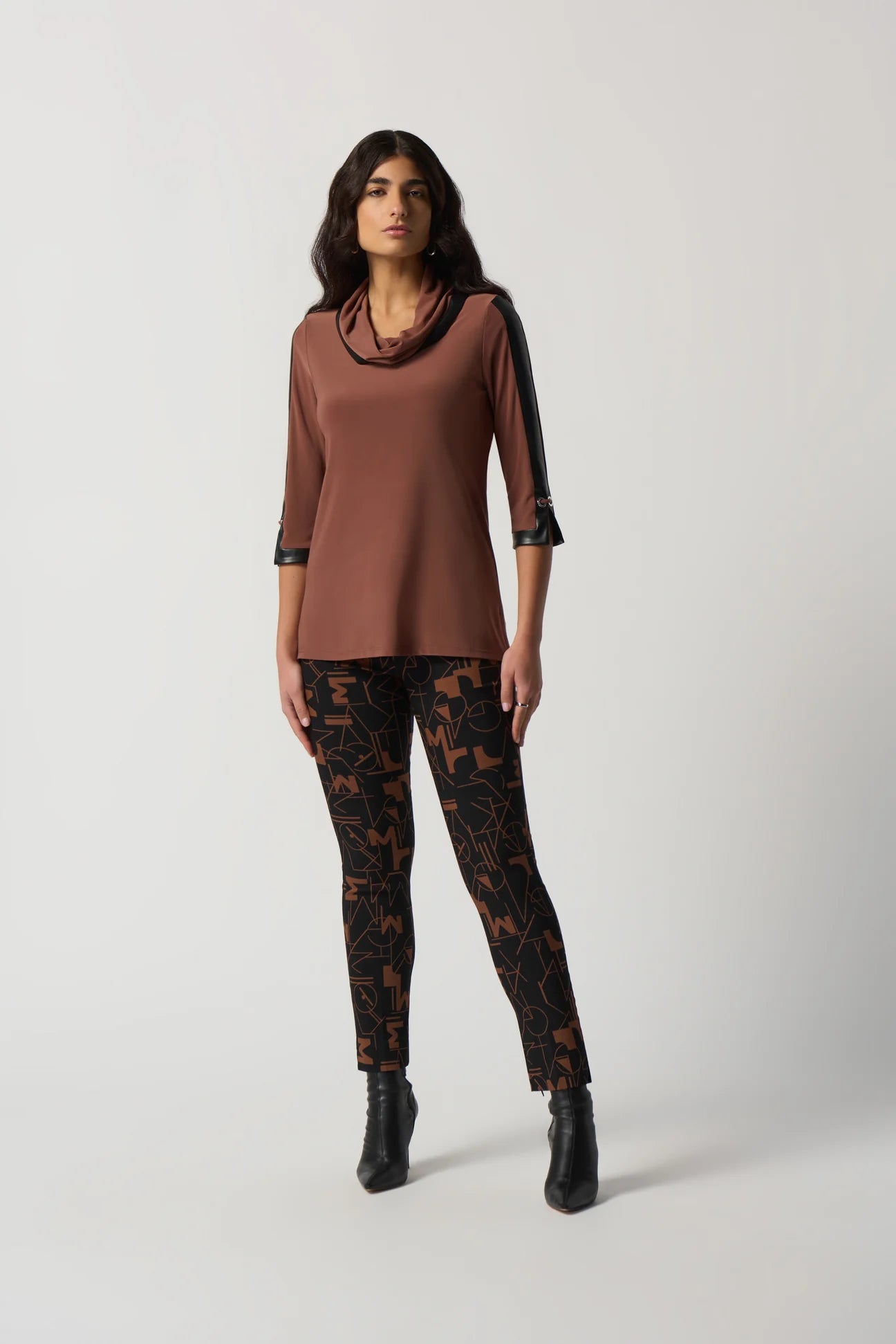 A woman with long dark hair is wearing the Joseph Ribkoff Toffee Faux-Leather Trim Tunic Style 233107, a brown long-sleeve top featuring black faux-leather trim on the shoulders and cuffs. She pairs it with black pants adorned with a geometric pattern. Standing against a plain white background, she gazes confidently at the camera.