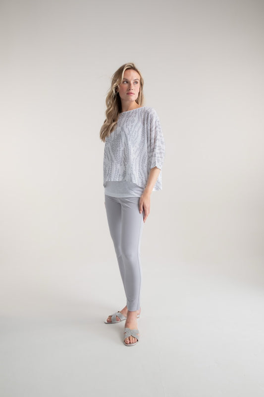 A woman with flowing blonde hair stands against a smooth, minimalistic backdrop. She wears MARBLE's oversized patterned top with tank top 7850 MARBLE, paired with light gray leggings and sandals, gazing slightly to the side.