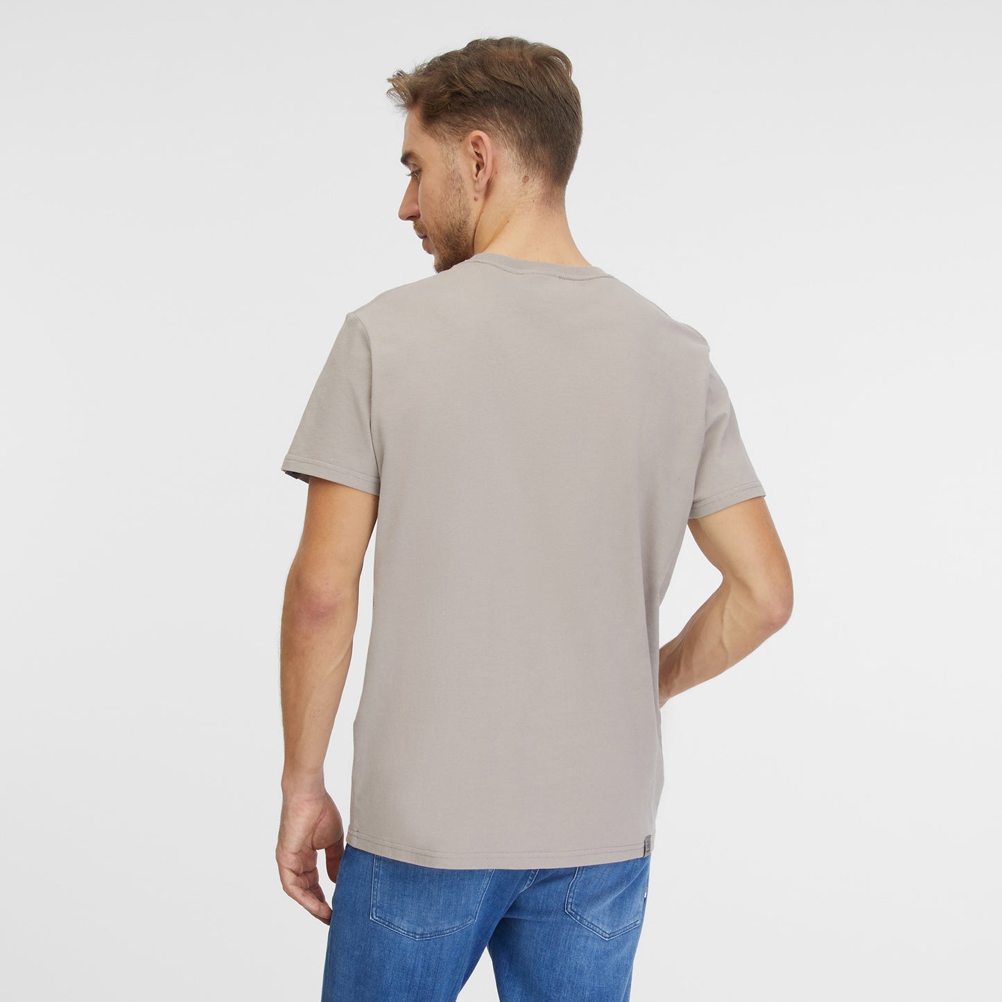 A man with short hair and stubble is wearing the Roggero T-Shirt 2422-15024-7001 from RAGWEAR, a beige, eco-friendly shirt featuring a mountain and bear graphic on the front. He pairs it with blue jeans and stands against a plain white background, looking directly at the camera with a neutral expression.