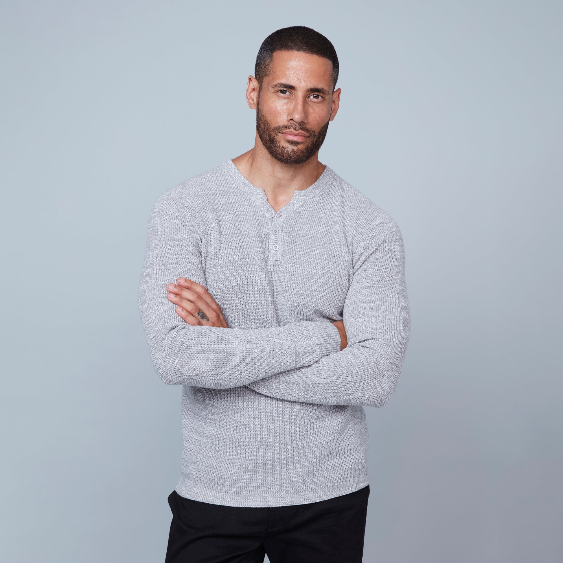 A stylish man with short hair and a beard stands against a grey background, looking down to his right with his arms crossed over his chest. He is wearing the light grey Henley Sweater Cotton from HEDGE, made from 100% cotton knit, paired with black pants.