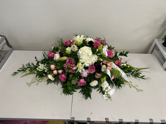 Introducing the "Pink & White - Casket Spray" by Posies Flowers & Fashion—a rectangular floral arrangement in a chic container featuring flowers like white hydrangeas, purple and pink roses, white lilies, and snapdragons. Accented with lush greenery and foliage, it is elegantly displayed on a white table against a plain wall. Please note that availability may vary seasonally.