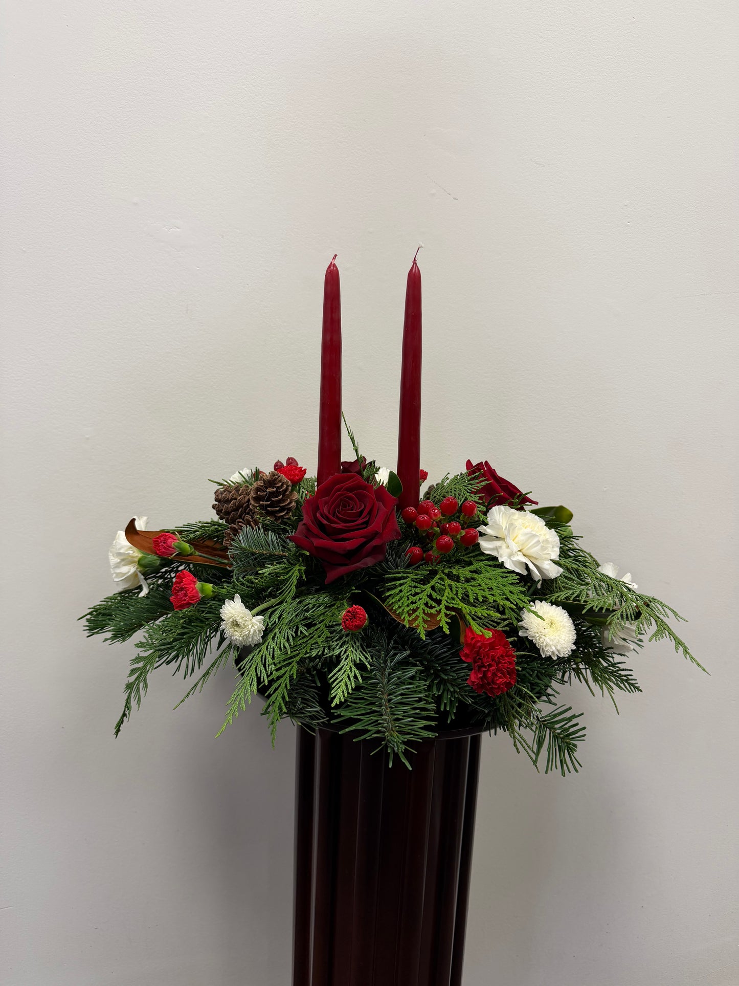 The Double Taper Candle Centrepiece with Roses by Posies Flowers & Fashion features red roses, white blooms, pinecones, greenery, and two tall red candles—ideal as a festive Christmas centrepiece against a white background.