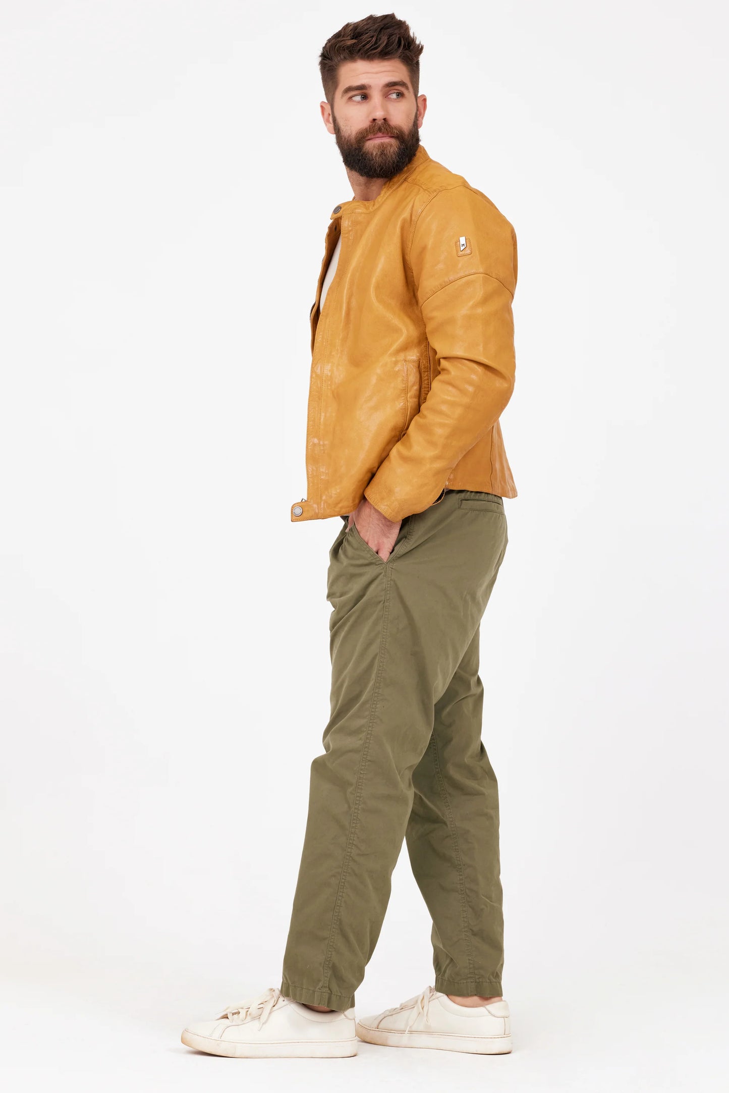 A bearded man is standing against a white background, wearing the MAURITIUS LEATHER Solvic CF Leather Jacket in Curry over a light gray T-shirt, accompanied by olive green cargo pants and white sneakers. His hands are in his pockets and he has a neutral expression on his face. The lightweight outfit contrasts sharply with the navy backdrop behind him.