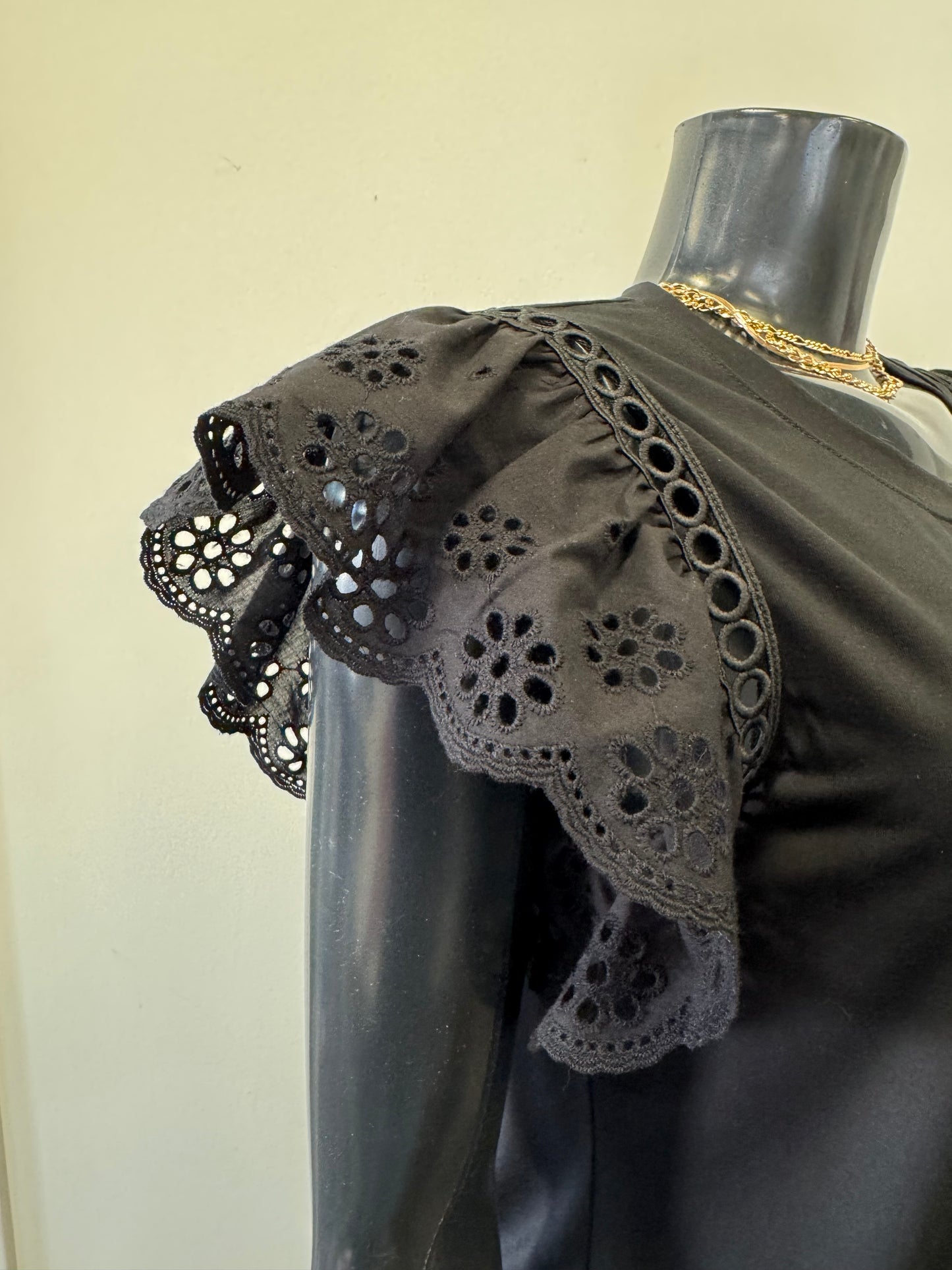 A black mannequin displays Top 8616 by Bali Corp, featuring short ruffled sleeves with lace eyelet details. Made from viscose and spandex, it ensures comfort. The mannequin is accessorized with a gold chain necklace.