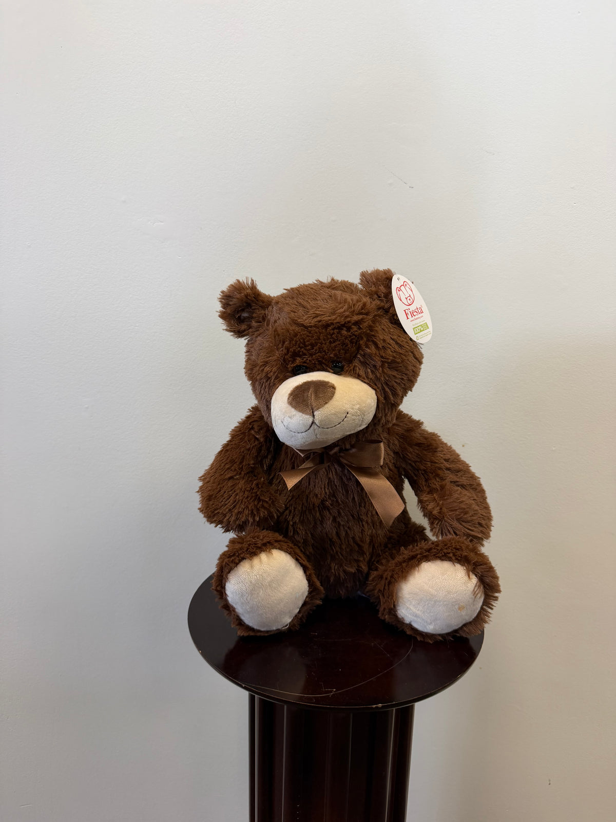 A Classic Teddy Bear - Brown 13 inch from Fiesta, featuring a white snout and paws with a ribbon and an ear tag, sits on a small round dark wooden table against a plain light-colored wall—making it an adorable choice for gift delivery.