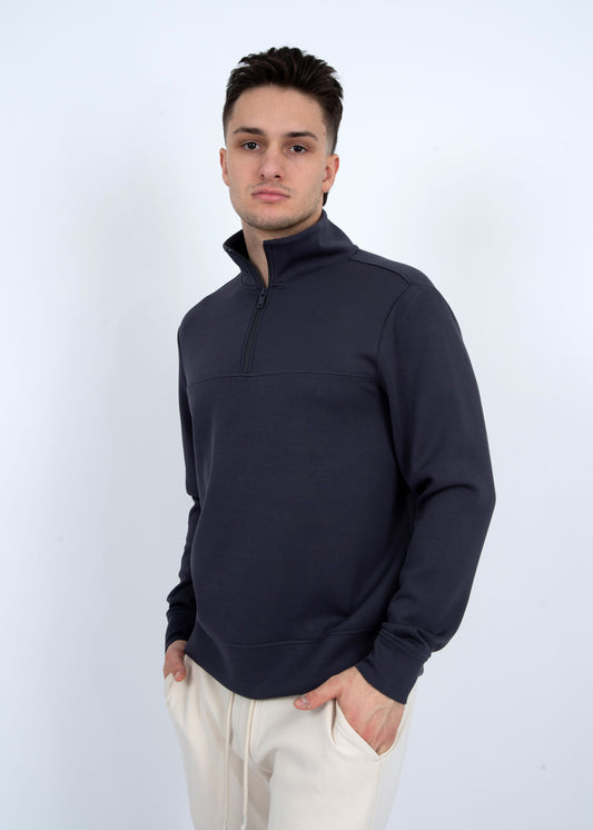 A person is against a plain background, wearing a soft knit pullover from HEDGE over a light blue 100% cotton shirt. With short hair and hands in pockets, they gaze directly at the camera.
