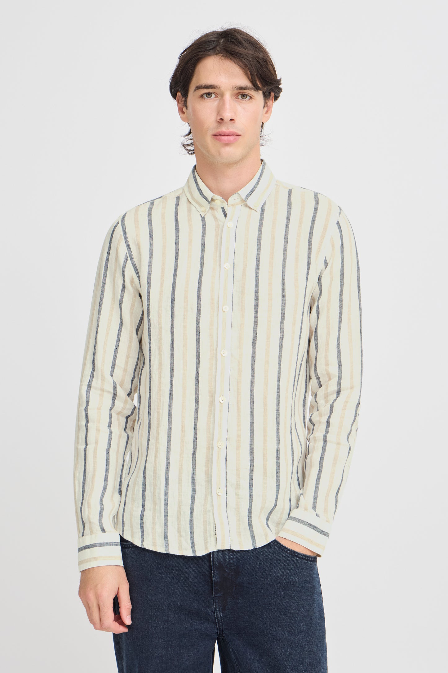 A person with long dark hair is casually dressed for the occasion in a Button Linen Cotton Shirt 20505463 by Casual Fridays, featuring white, beige, and blue vertical stripes. They stand with one hand in their dark jeans pocket against a plain white background.