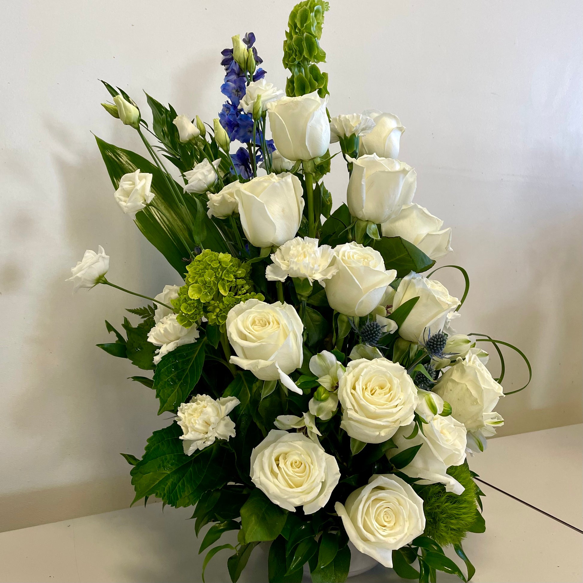 The "Pure Serenity" arrangement by Posies Flowers & Fashion offers an elegant sympathy tribute, showcasing a mix of white roses, white lilies, and green leaves beautifully presented in a white ceramic vase against a plain background.