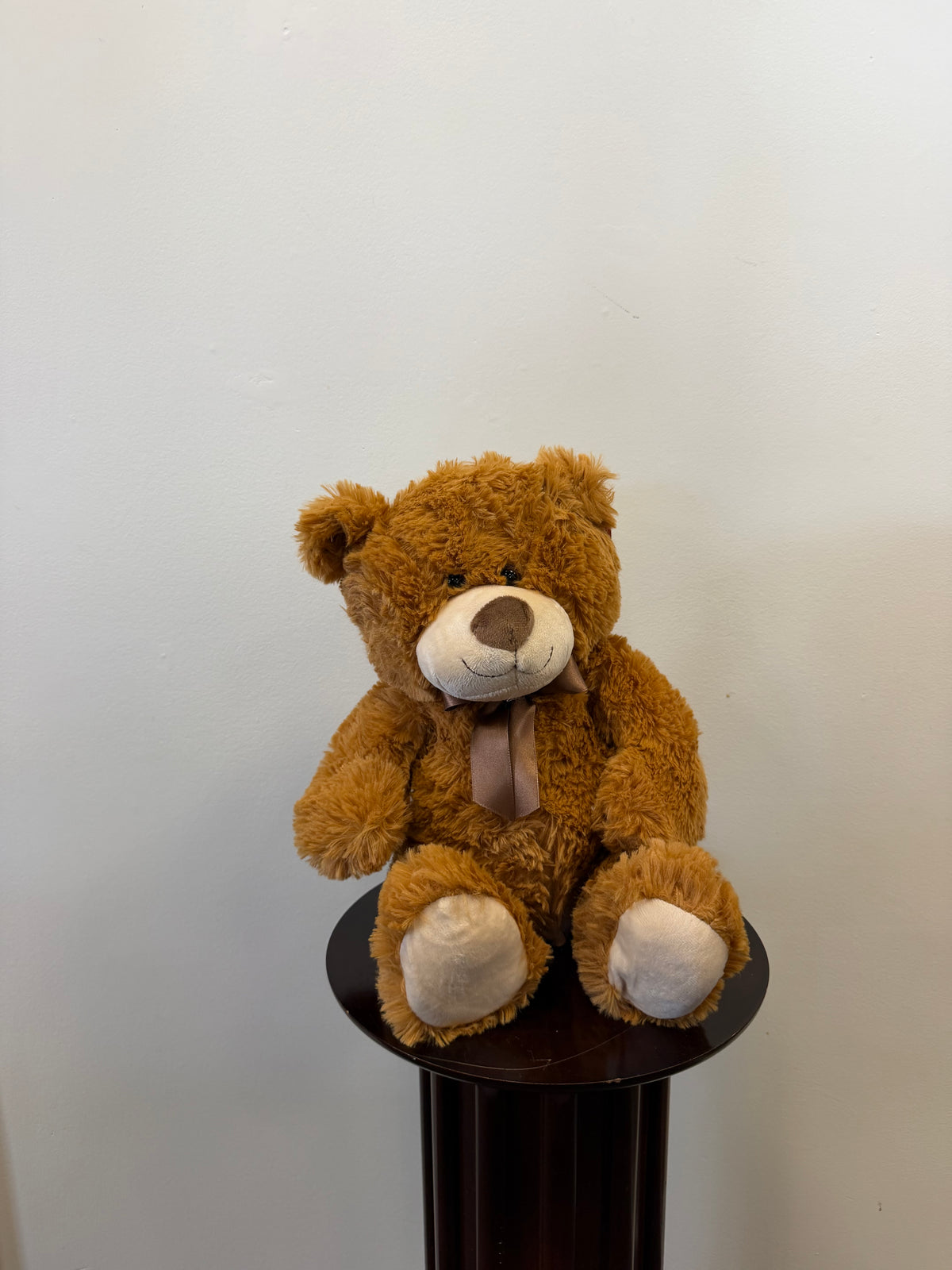 The Classic Teddy Bear - Light Brown 13 inch by Fiesta sits on a small round dark stand against a plain white wall. This plush bear from Fiesta features a beige snout and paws and is adorned with a light brown ribbon around its neck, enhancing its timeless charm.