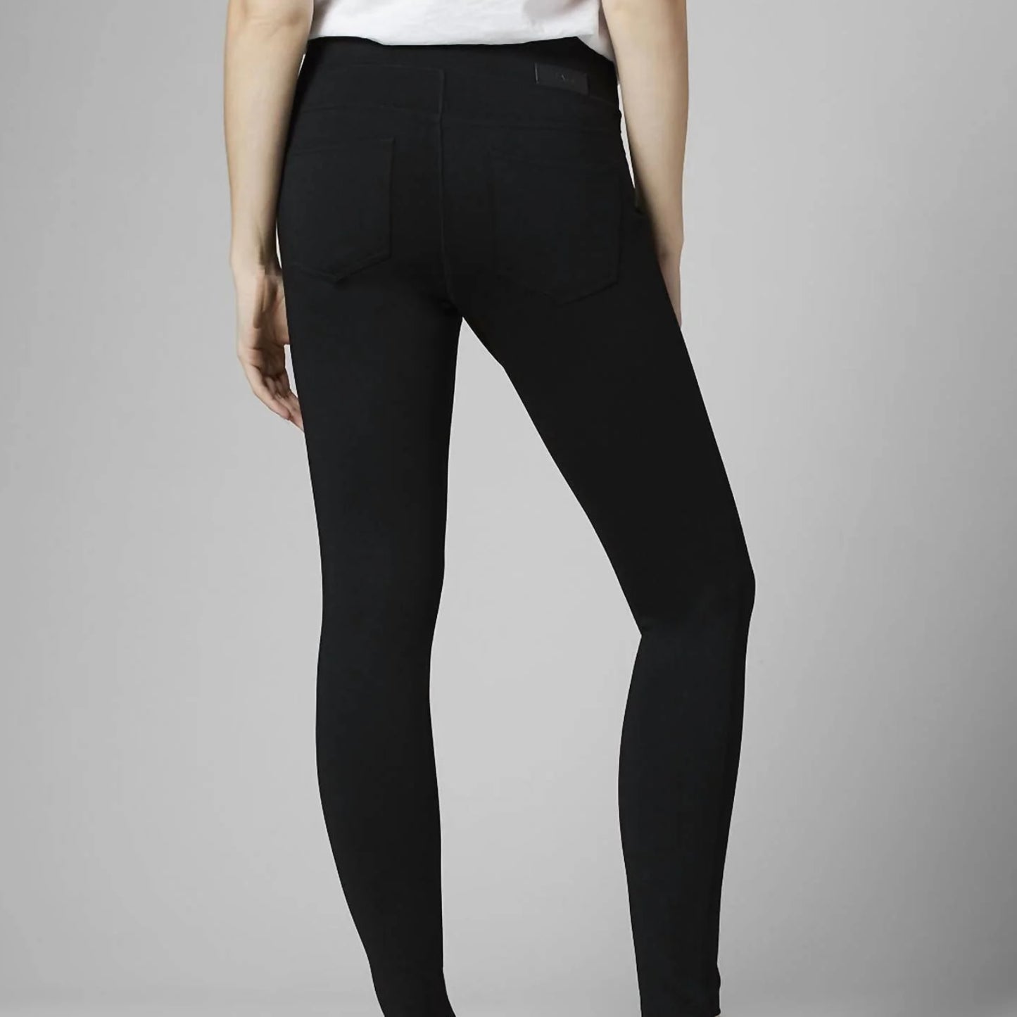 A woman stands against a plain gray background, dressed casually in a white short-sleeve T-shirt with a pocket on the left side. She is wearing the Ricki Mid Rise Leggings in Black by JAG, which are made from power stretch fabric. With one hand by her side and the other slightly in her pocket, she conveys a relaxed pose.