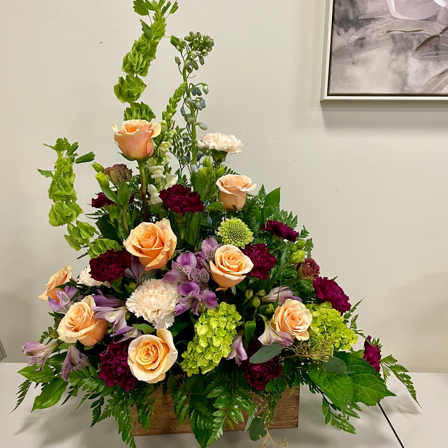 Introducing the Gentle Memories arrangement by Posies Flowers & Fashion—a lush funeral tribute featuring peach roses, green hydrangeas, purple alstroemeria, burgundy carnations, and white chrysanthemums. Accented with greenery, it creates a comforting atmosphere against a plain white background.
