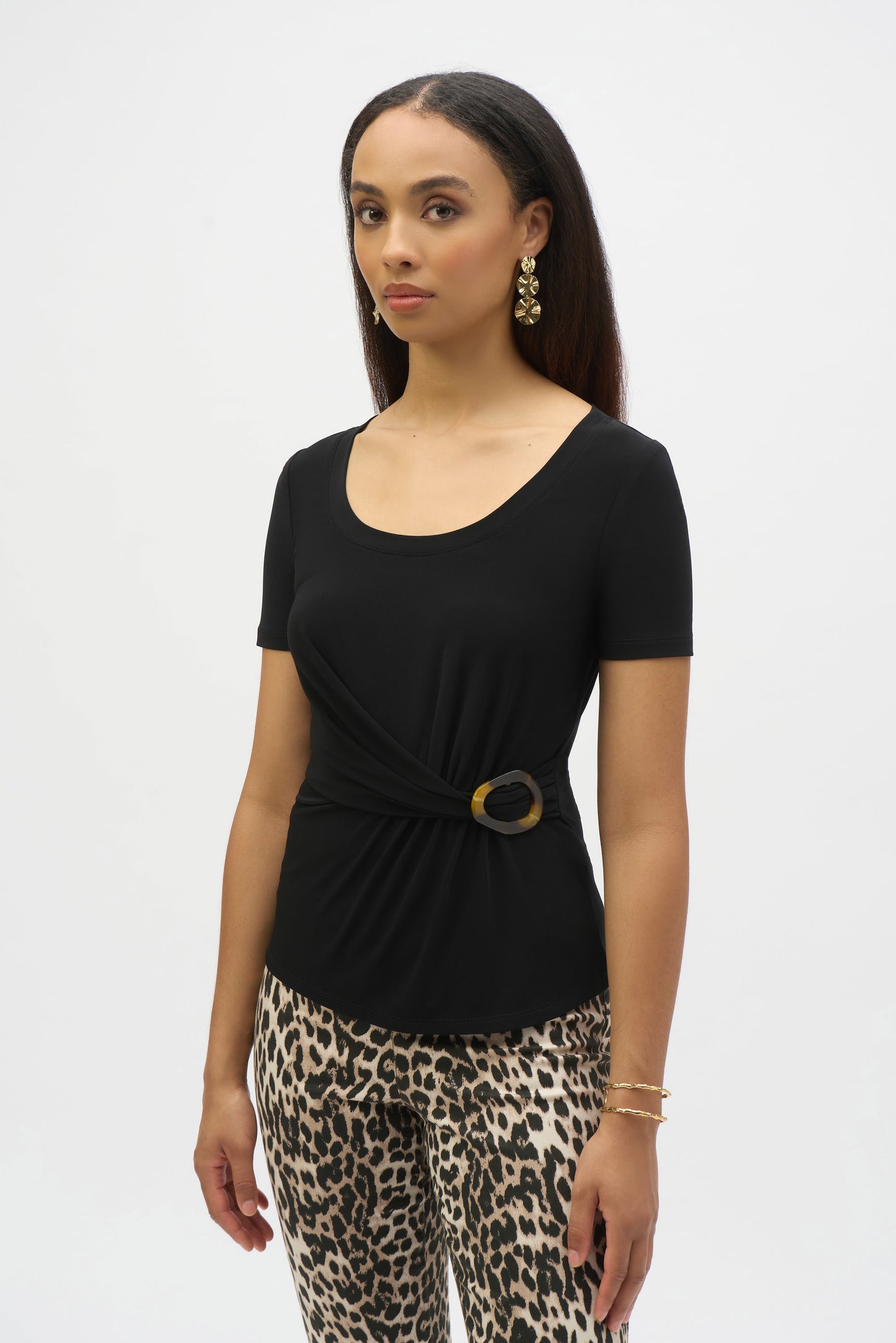 A woman with long hair wears a Joseph Ribkoff Silky Knit Fitted Top With Buckle Detail 252230, paired with leopard print pants. She accessorizes with gold dangling earrings and bracelets, standing against a plain background.