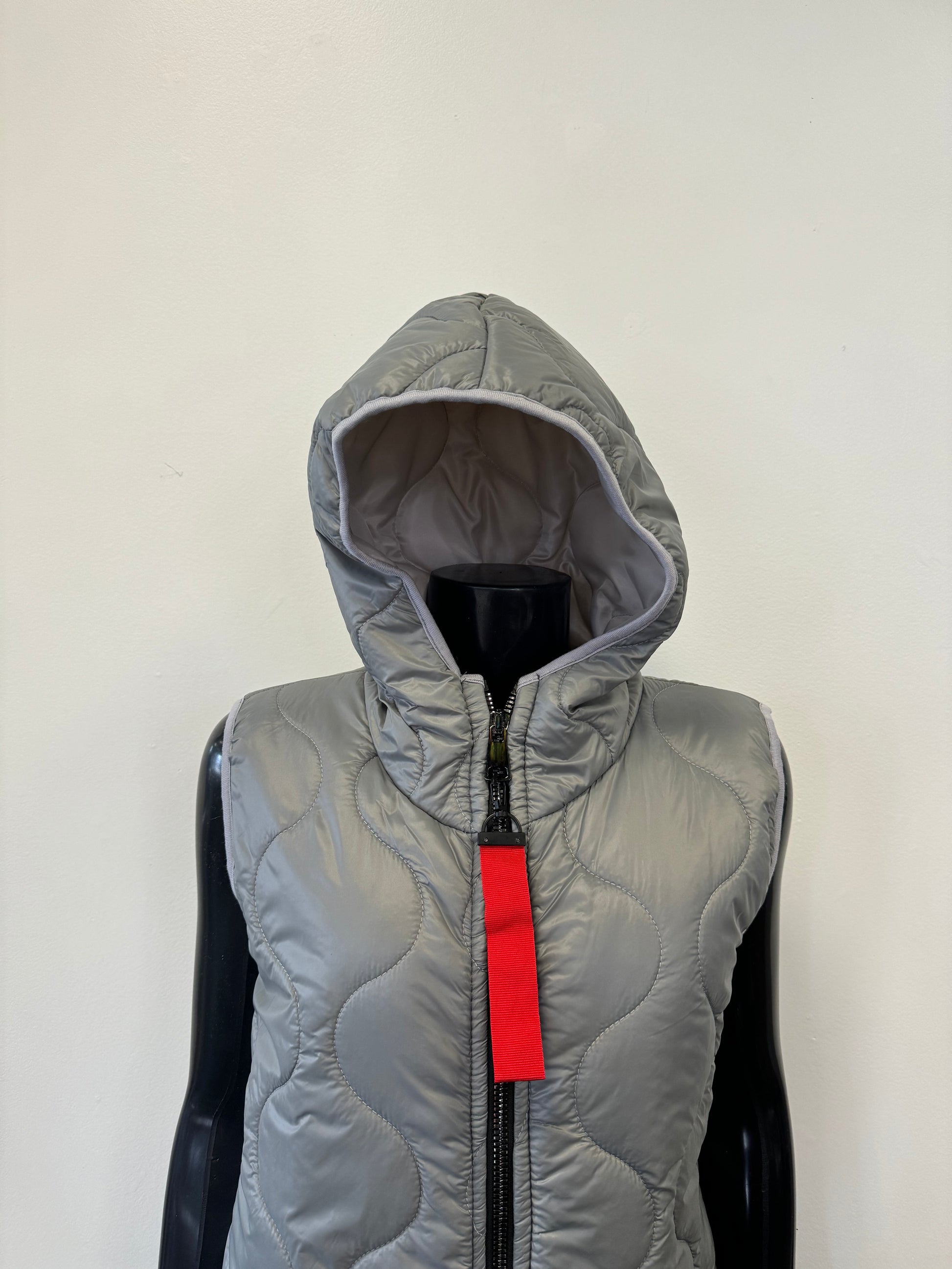 A black mannequin showcases the Vest Light by Catherine Lillywhite, a chic sleeveless and hooded gray quilted piece. The stylish vest features a distinctive wavy stitch pattern and a front zipper adorned with a red tag. An off-white wall serves as a simple backdrop for the display.