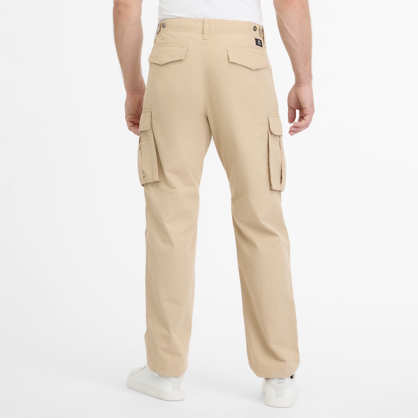 A person is seen from the rear wearing RAGWEAR's vegan streetwear, specifically the MERLY PANTS 2512-55004-6018. These ethical beige cargo pants have large side pockets with button flaps, complemented by white sneakers, set against a plain white background.