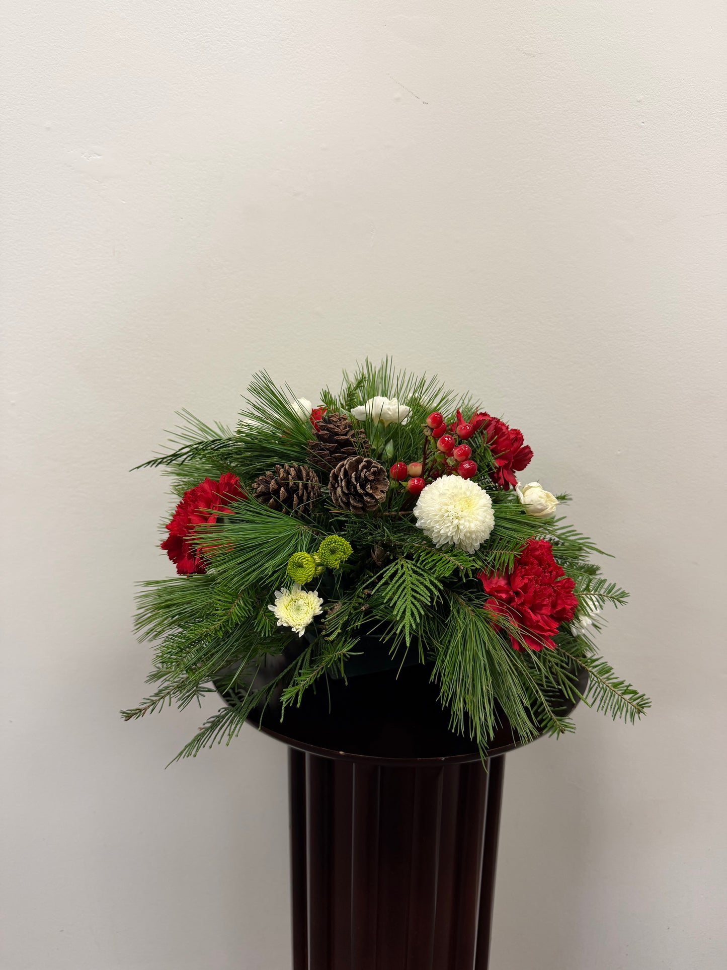 The "More Simple Christmas" arrangement by Posies Flowers & Fashion features red and white flowers, pine cones, greenery, and evergreens on a dark pedestal, adding festive cheer to any space.