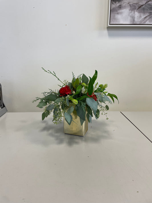 The "Designer’s Choice Centrepiece" by Posies Flowers & Fashion is a small floral arrangement with red and green foliage in a square, light-colored vase on a gray surface, set against a white wall with a partially visible framed picture.