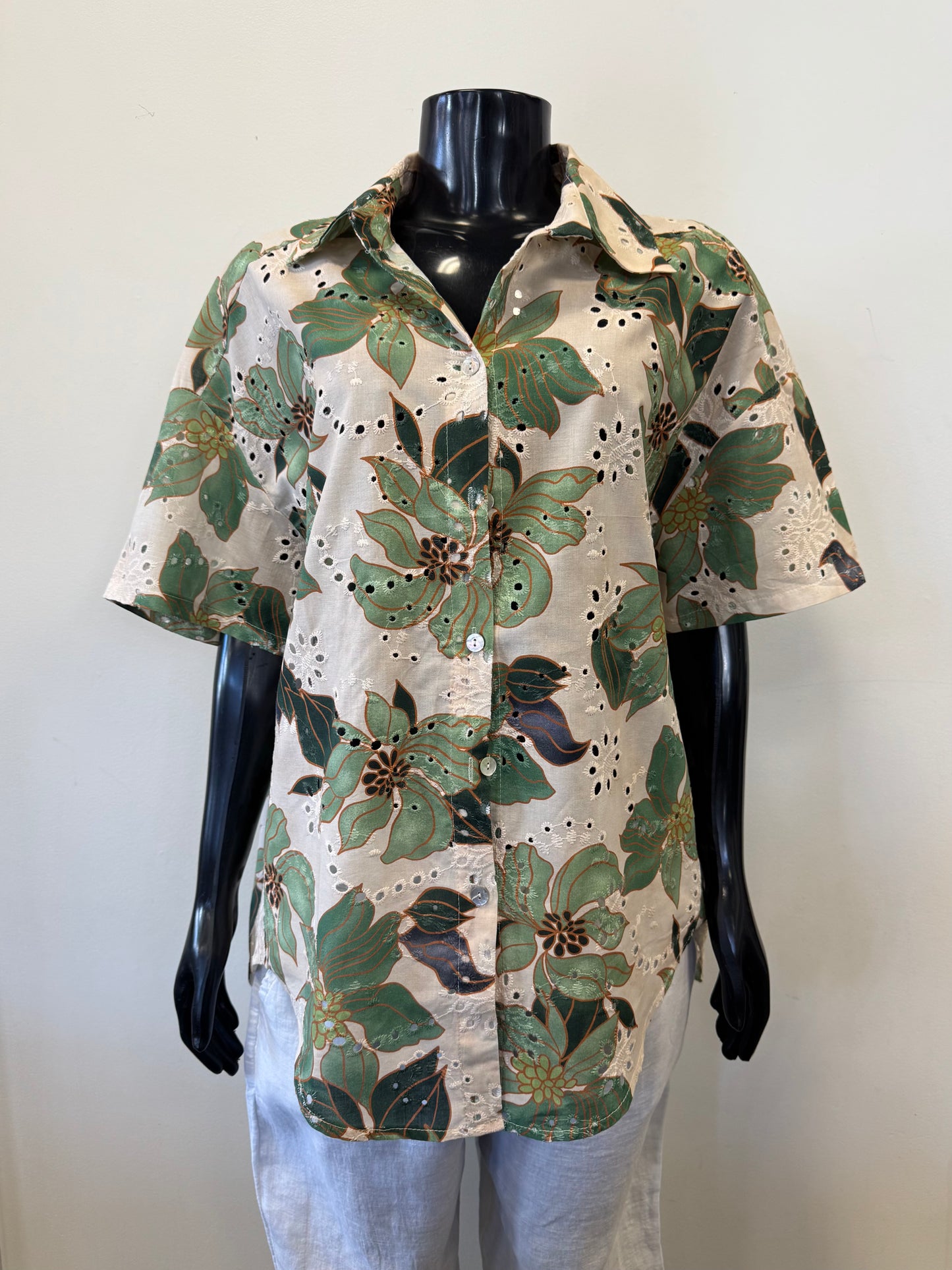 The Top Cotton 877 from Bella Amore, a luxurious floral button-up crafted from exquisite Italian cotton, showcases green leaves and large flowers on a black mannequin. With its short sleeves and eyelet details, it pairs perfectly with light-colored linen pants.