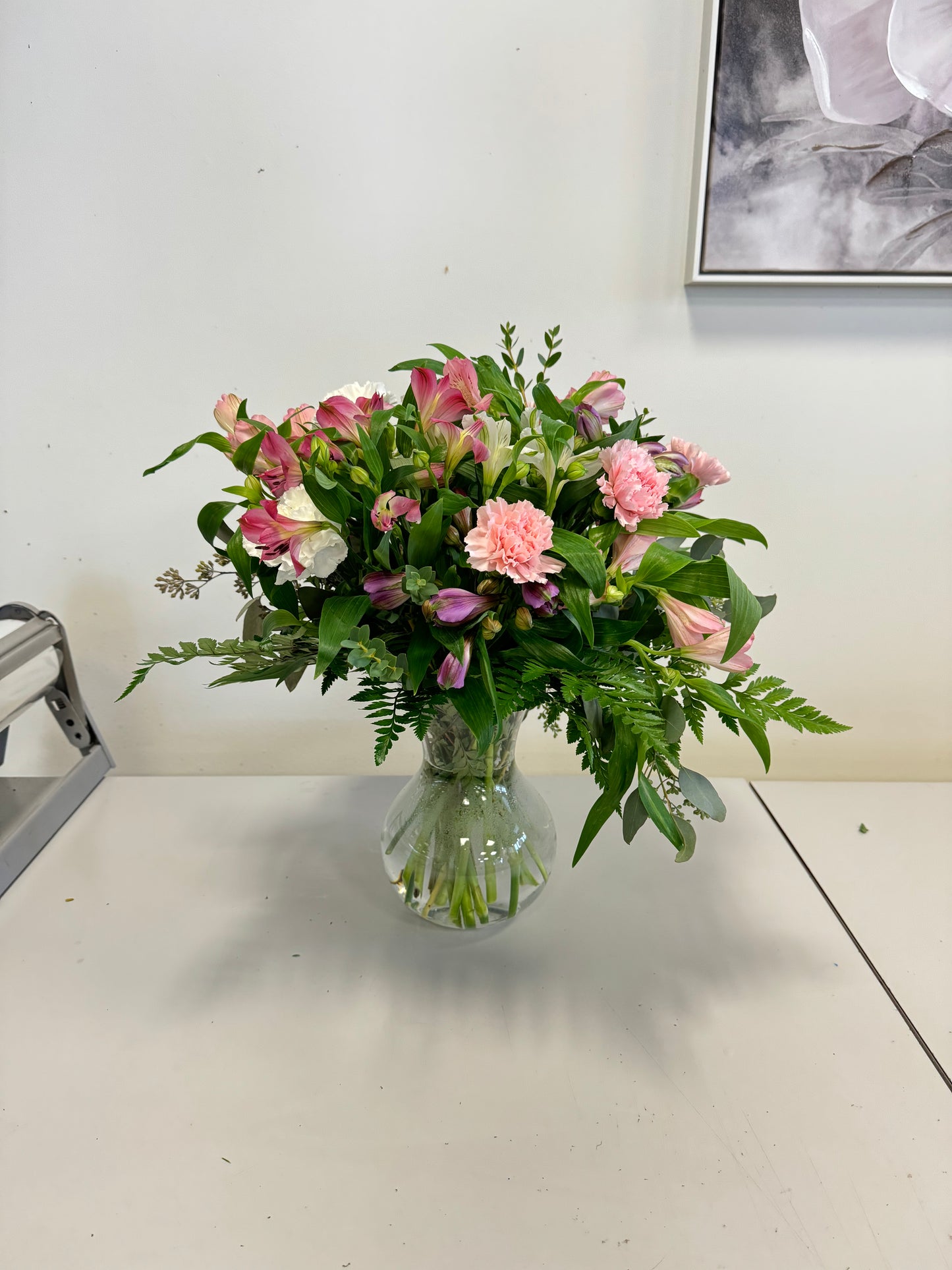 A Designer’s Choice Vase from Posies Flowers & Fashion, filled with white lilies and green foliage, graces a marble table in an elegantly modern room. Soft lighting and contemporary furniture in the background create a warm and inviting atmosphere, showcasing the artistry of floral designers, subject to availability.