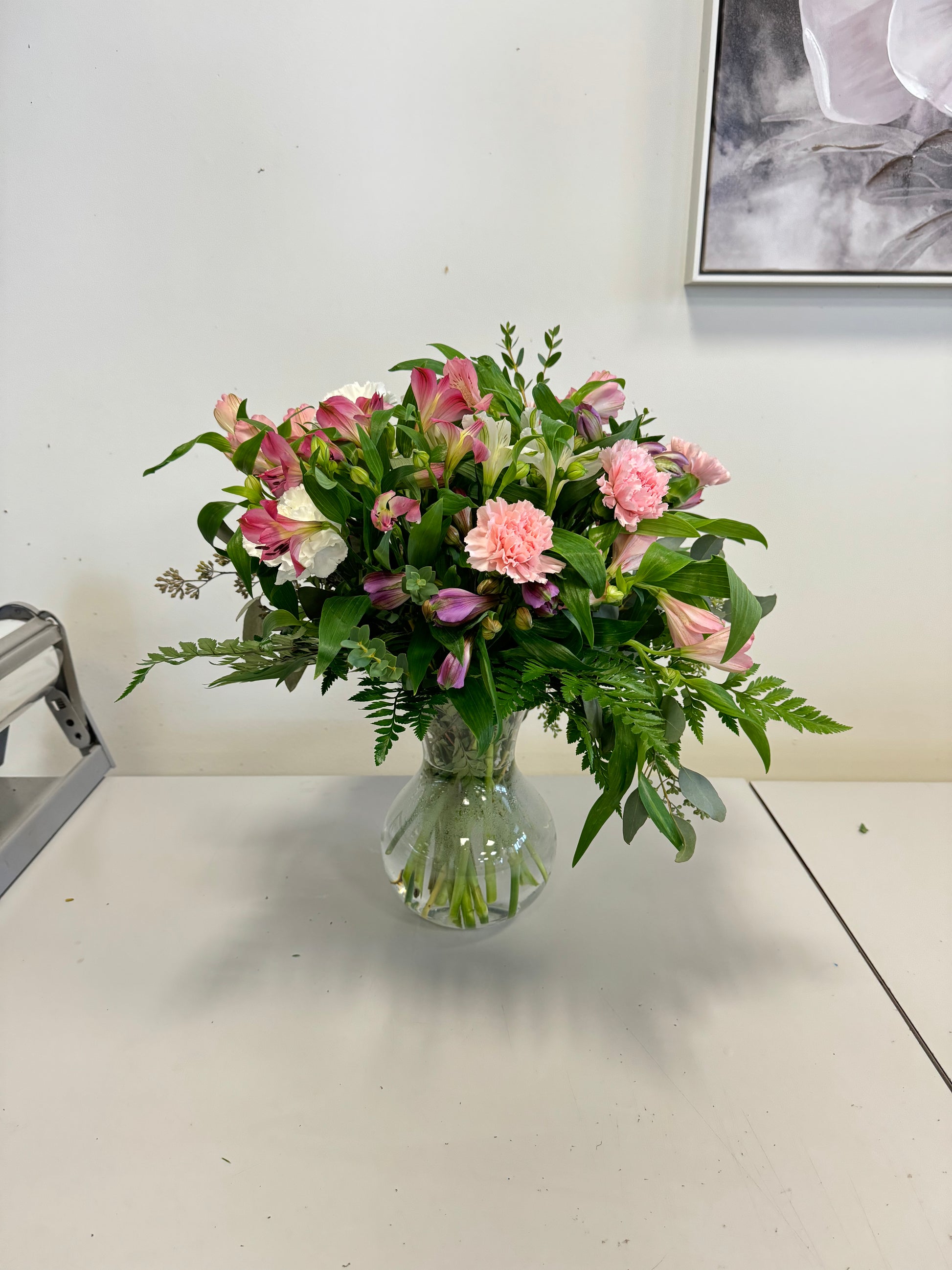 A Designer’s Choice Vase from Posies Flowers & Fashion, filled with white lilies and green foliage, graces a marble table in an elegantly modern room. Soft lighting and contemporary furniture in the background create a warm and inviting atmosphere, showcasing the artistry of floral designers, subject to availability.