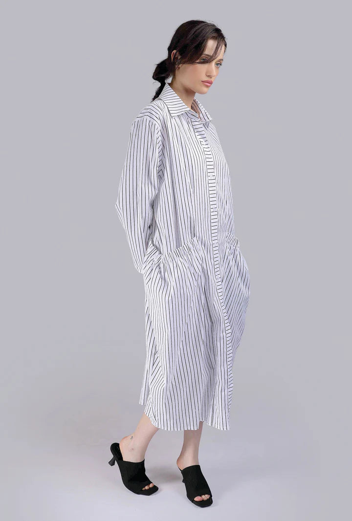A woman in profile wears the Paolo Tricot Dress VT-3037—a striped Italian cotton shirt dress with long sleeves and pockets. Her dark hair is tied back, and she pairs it with black heeled sandals against a plain light background.