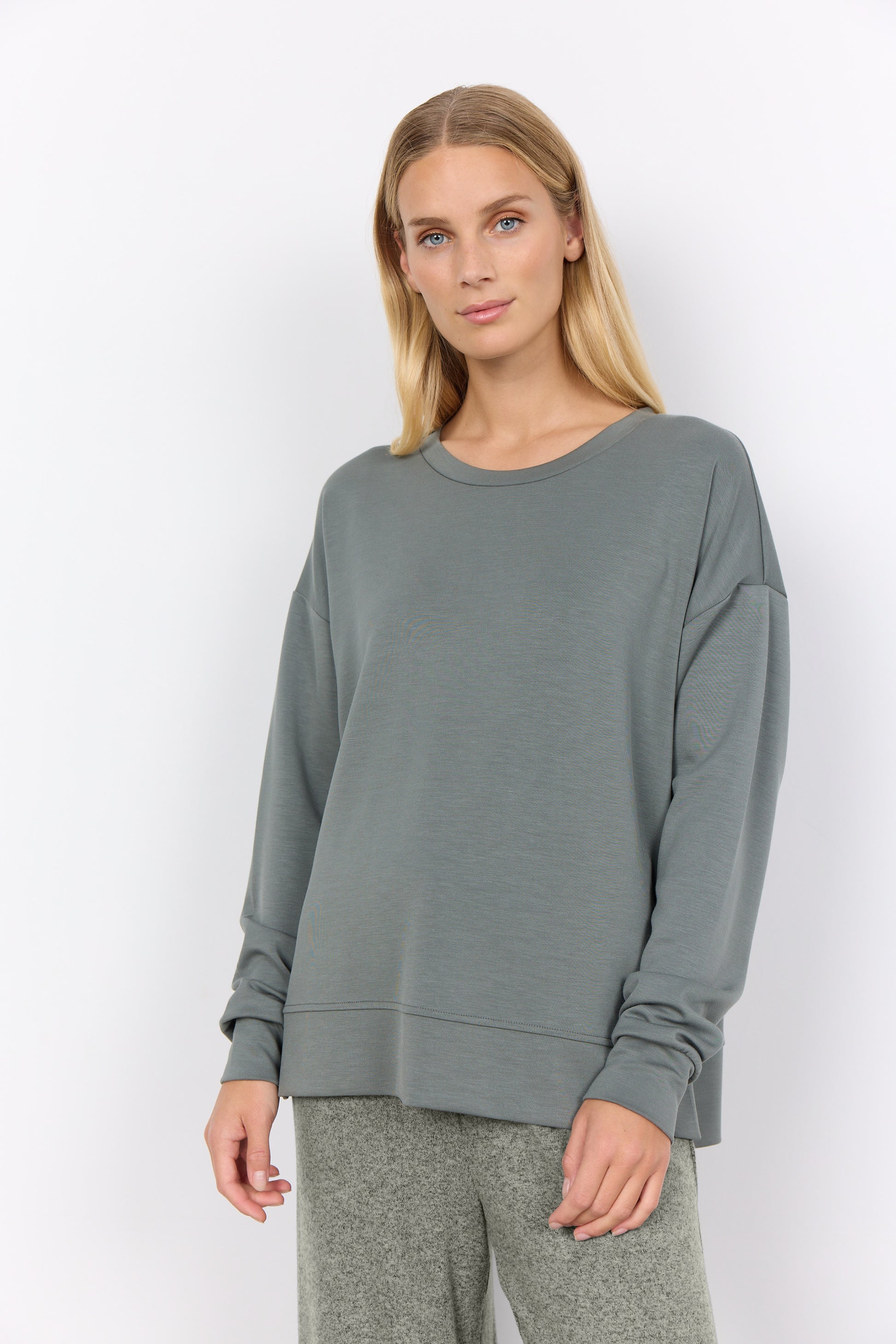 The woman elegantly poses against a white backdrop, wearing the trendy Sweatshirt Banu 164 SoyaConcept with matching gray pants. Her relaxed posture and gentle smile enhance the effortlessly chic ensemble, making it a perfect wardrobe addition.