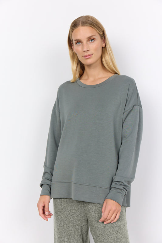 The woman elegantly poses against a white backdrop, wearing the trendy Sweatshirt Banu 164 SoyaConcept with matching gray pants. Her relaxed posture and gentle smile enhance the effortlessly chic ensemble, making it a perfect wardrobe addition.