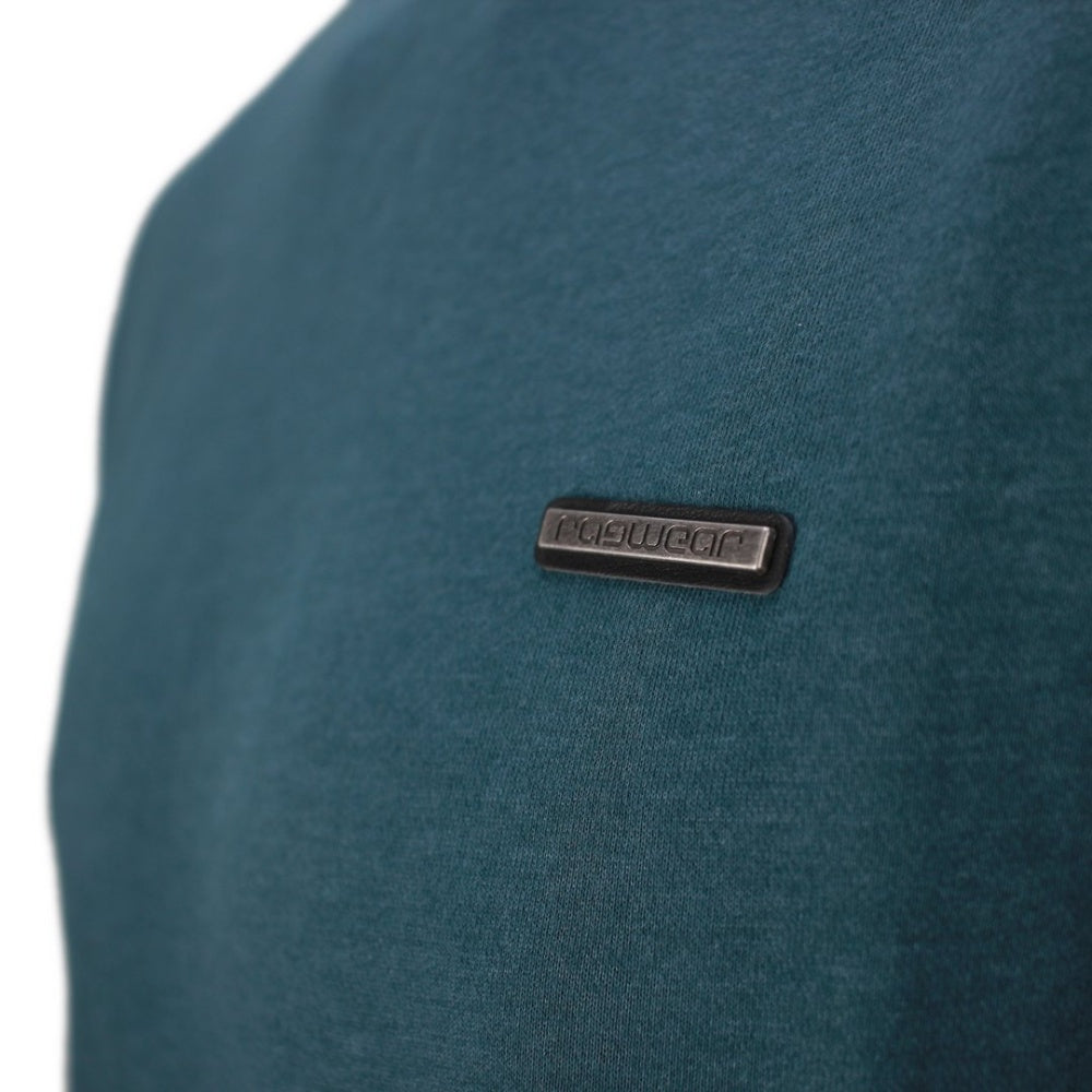 A smiling man with short brown hair is wearing a teal Ragwear Sweatshirt Pullover - INDDIE from RAGWEAR and black pants. He is standing against a plain white background, slightly turned to the side with his hands relaxed by his sides, showcasing a casual yet sporty look.