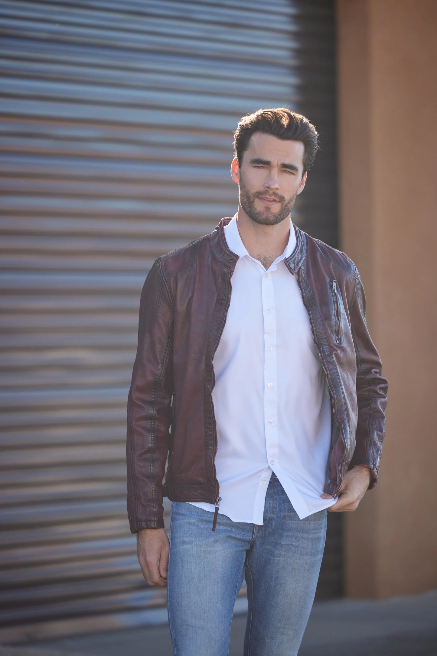 A man with dark hair and a beard stands in front of a corrugated metal background, wearing a white dress shirt and a premium quality MAURITIUS LEATHER Rakva RF Leather Jacket in wine, with his hands in his jacket pockets.
