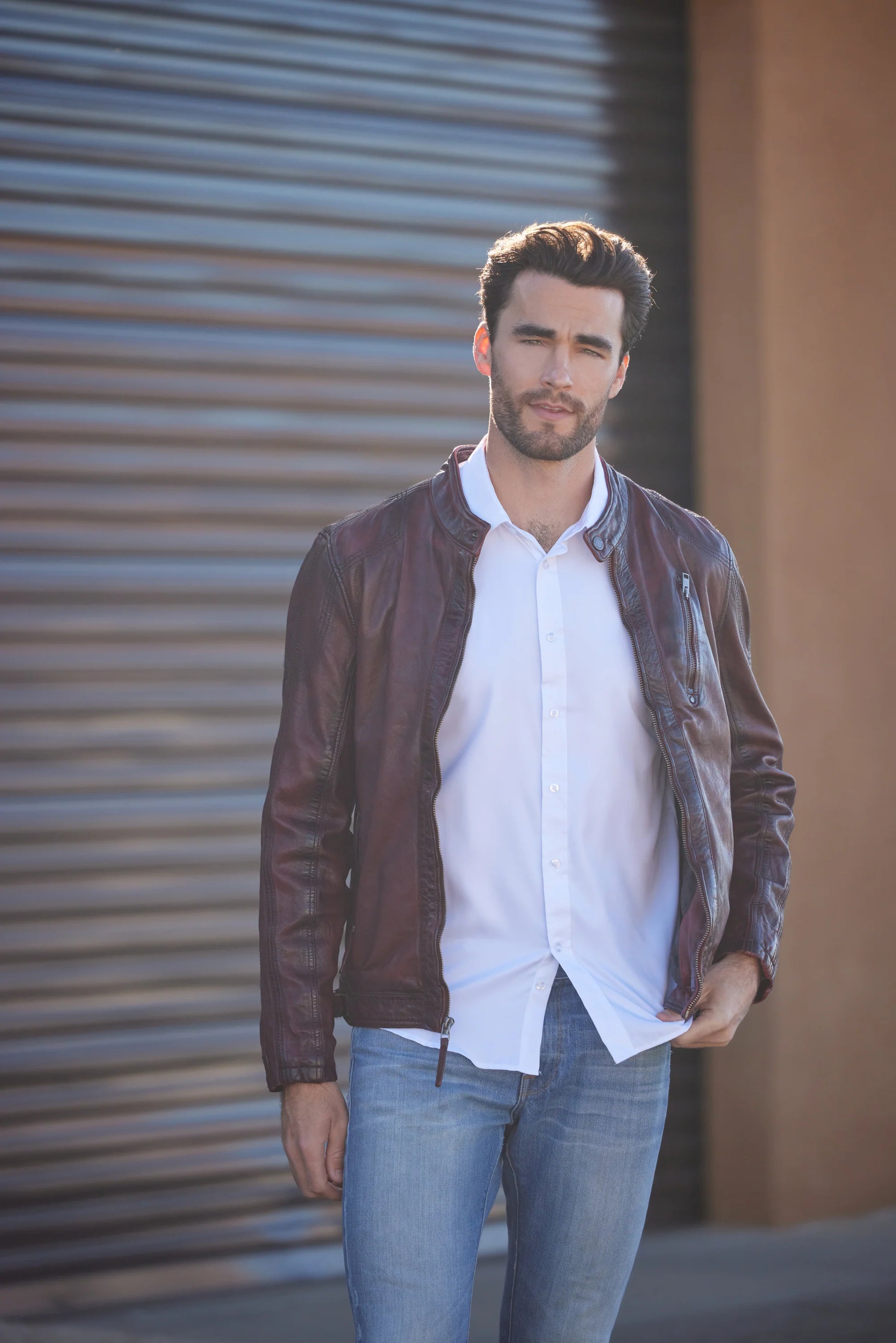 A man with dark hair and a beard stands in front of a corrugated metal background, wearing a white dress shirt and a premium quality MAURITIUS LEATHER Rakva RF Leather Jacket in wine, with his hands in his jacket pockets.