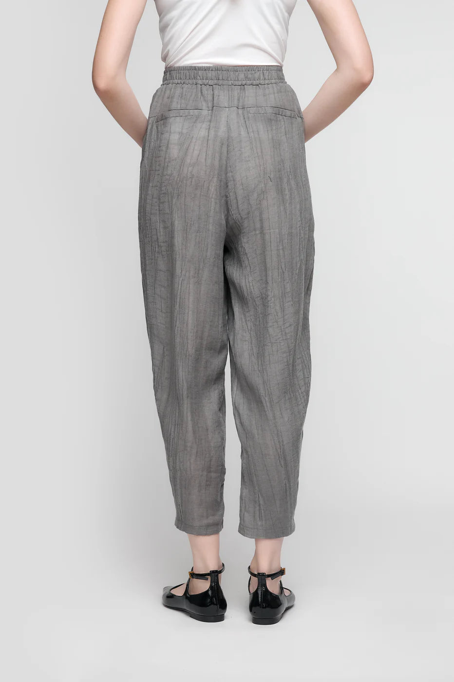 A person stands with their back to the camera in a white top and likely PANTS WITH FRONT POCKETS AND ELASTIC WAIST - ANTHRACITE- CS25-116 by UCHUU, complemented by black ankle-strap flats against a light gray background.
