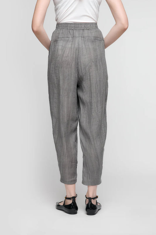 A person stands with their back to the camera in a white top and likely PANTS WITH FRONT POCKETS AND ELASTIC WAIST - ANTHRACITE- CS25-116 by UCHUU, complemented by black ankle-strap flats against a light gray background.