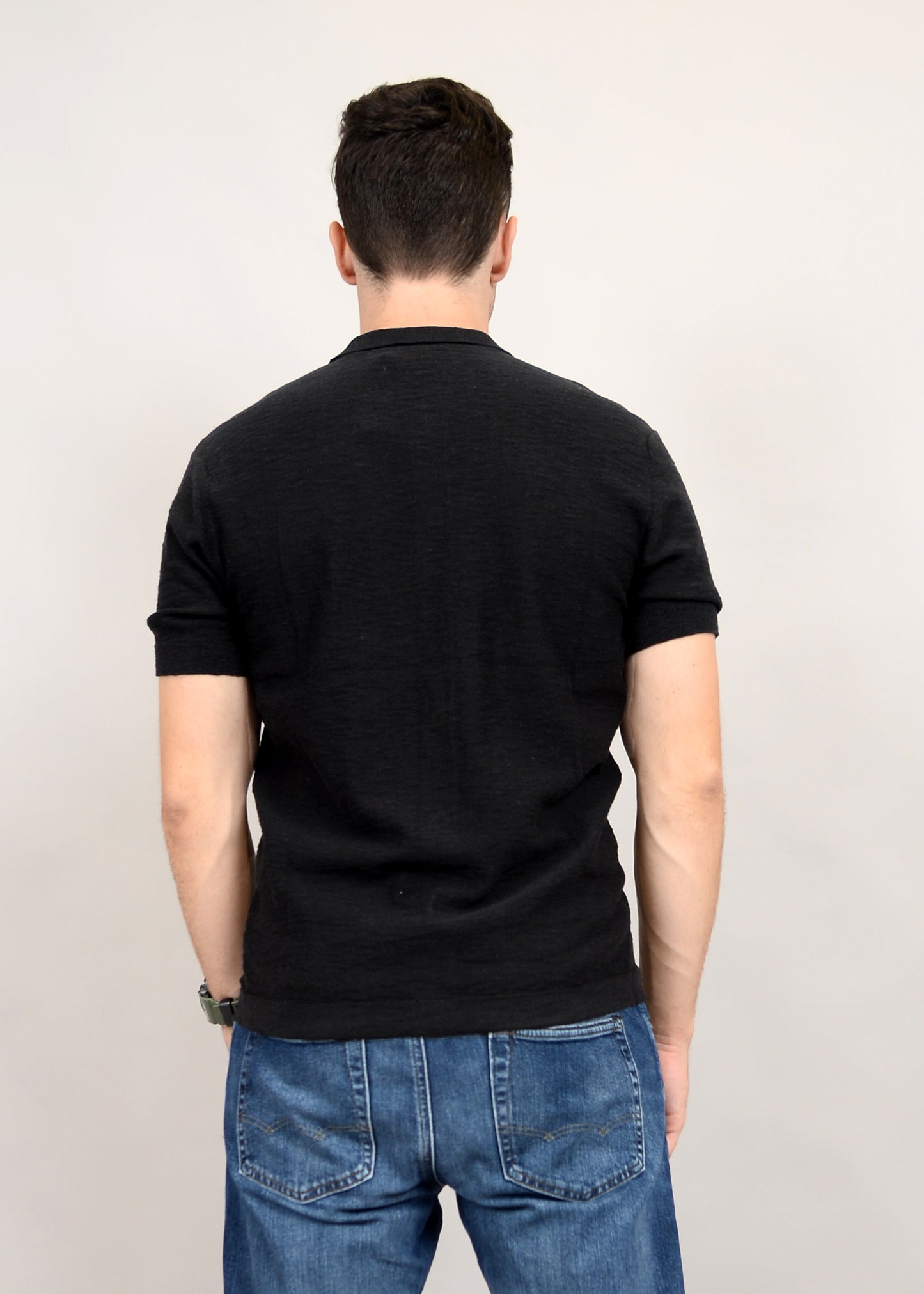 Against a plain background, a man stands with his back to the camera in fashionable blue jeans and a black T-shirt, suggesting a versatile wardrobe featuring the HEDGE Sweater Knit Button Shirt Short Sleeve 56MS311S made from 100% cotton.