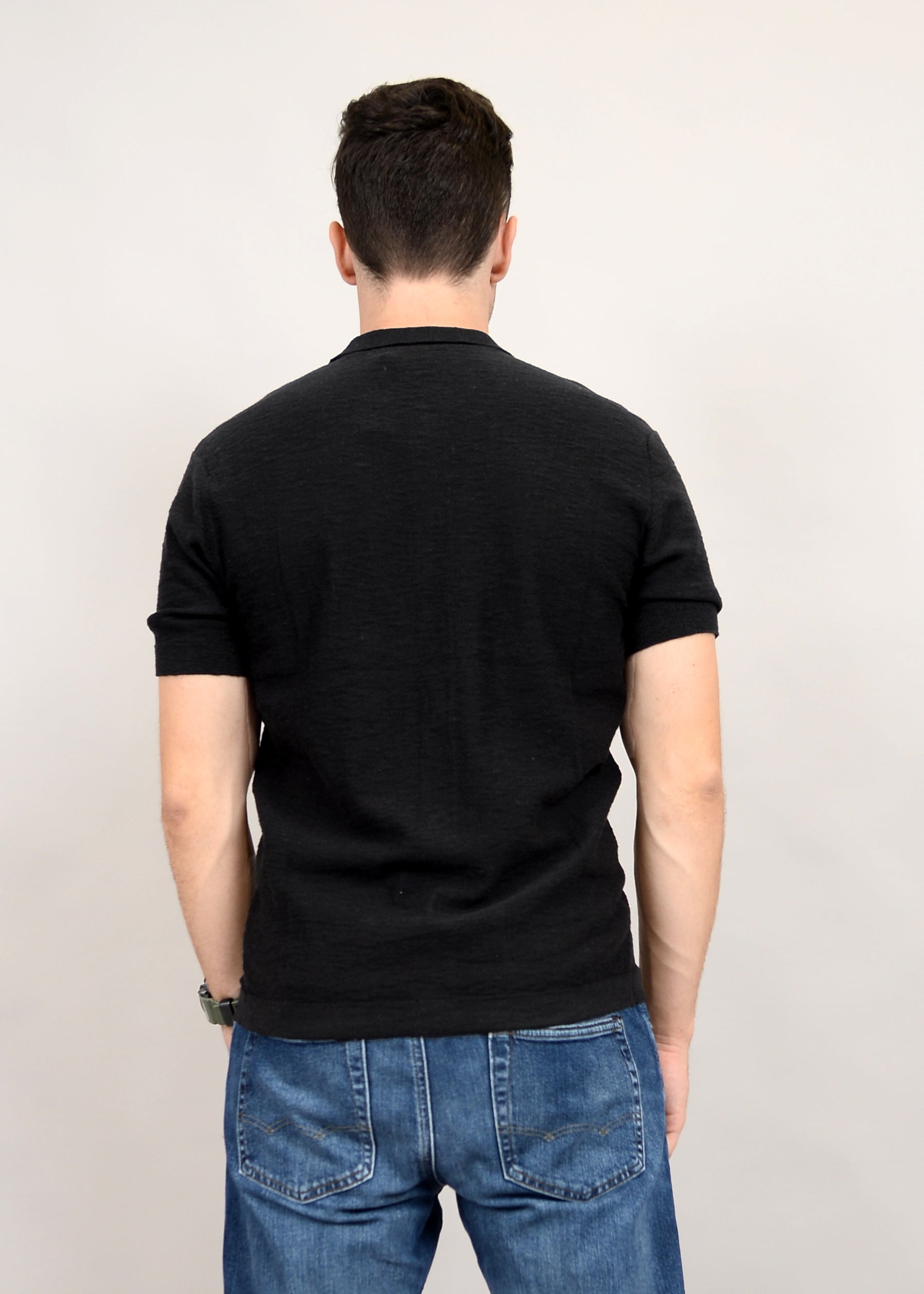 Against a plain background, a man stands with his back to the camera in fashionable blue jeans and a black T-shirt, suggesting a versatile wardrobe featuring the HEDGE Sweater Knit Button Shirt Short Sleeve 56MS311S made from 100% cotton.