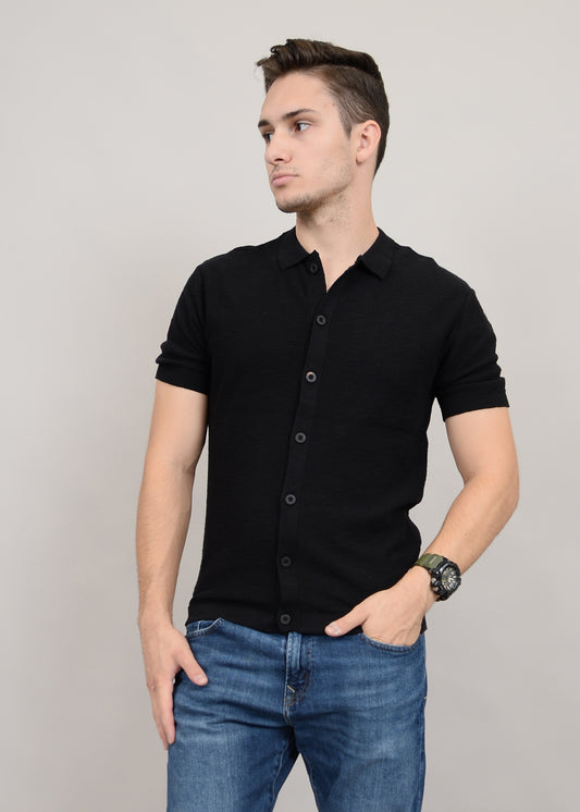A person with short hair stands against a plain background, looking to the side in a fashion-forward wardrobe, wearing a sleek HEDGE Sweater Knit Button Shirt Short Sleeve 56MS311S and blue jeans with a watch on their left wrist, effortlessly merging style with simplicity.