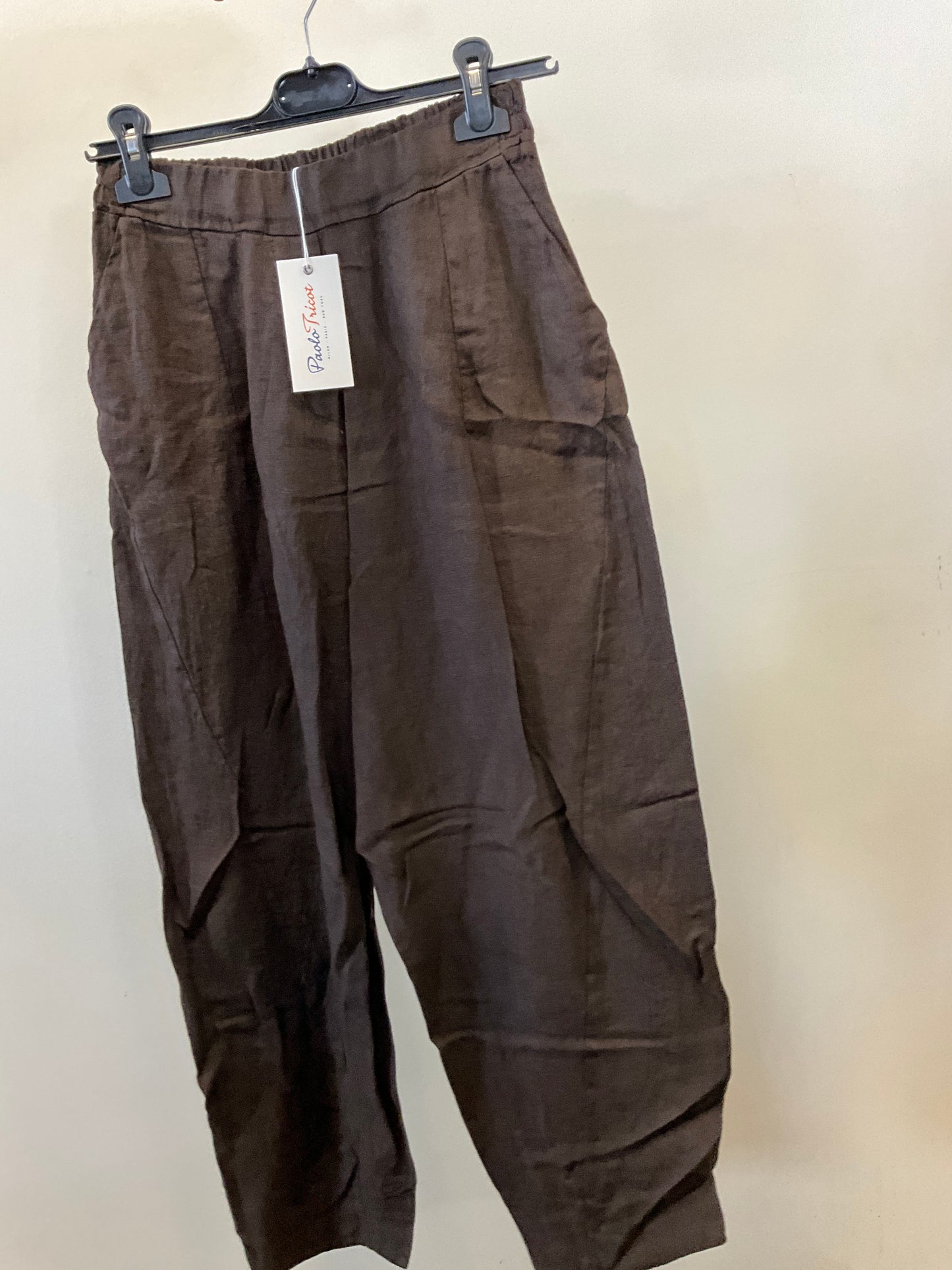 The Pant 100% Linen Chocolate by Paolo Tricot, featuring a loose fit and elastic waistband, hangs on a black hanger with a white tag attached. The chocolate brown pants are displayed against a plain off-white wall.