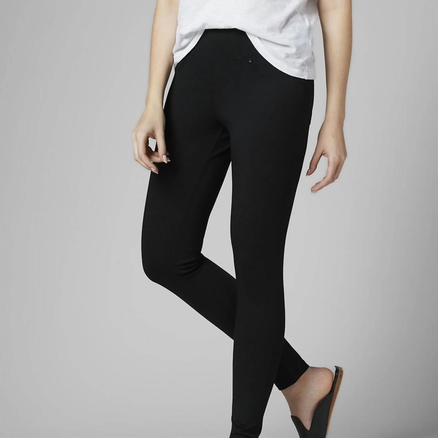 A woman stands against a plain gray background, dressed casually in a white short-sleeve T-shirt with a pocket on the left side. She is wearing the Ricki Mid Rise Leggings in Black by JAG, which are made from power stretch fabric. With one hand by her side and the other slightly in her pocket, she conveys a relaxed pose.