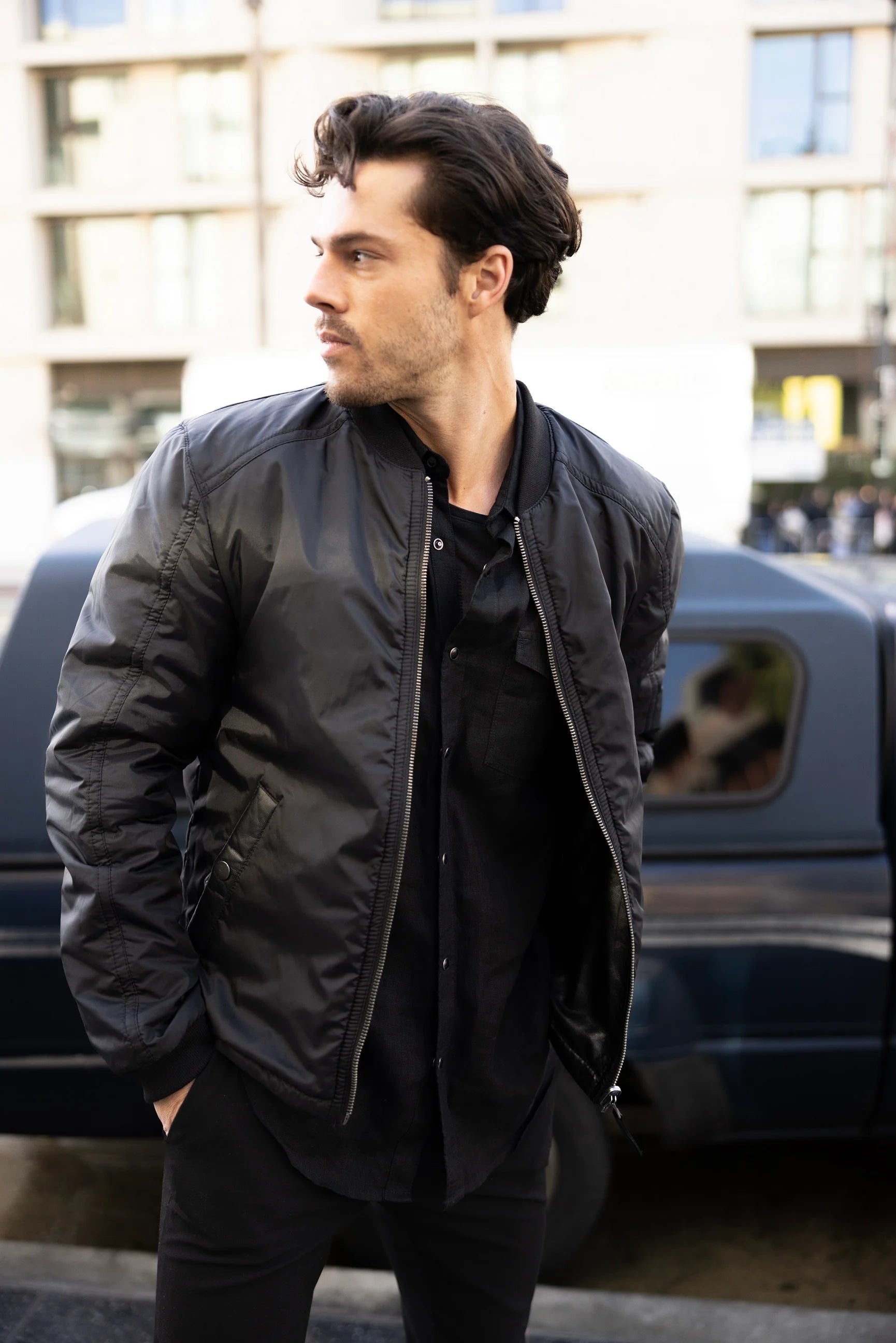 A person with dark, wavy hair and facial hair stands against a brick wall. They are wearing a Cranch CF Leather Jacket, Black - Reversible by MAURITIUS LEATHER, made from lambskin leather over a black shirt, with one hand in the jacket pocket and maintaining a neutral expression.