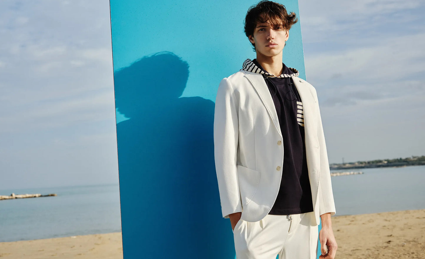 A man stands on a beach next to a large blue square panel, wearing the Paul Miranda Blazer Panna GB1374JK and matching trousers over a black and white striped shirt. His shadow is cast on the panel, and the sea is visible in the background under a cloudy sky—a perfect scene for a special occasion.