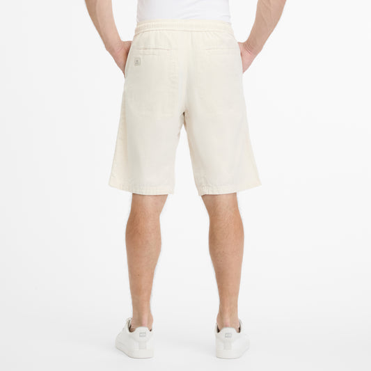 A person is seen from the back wearing RAGWEAR's LONGYZ 2512-50014-7010 light beige shorts made of a cotton linen blend with two back pockets, paired with white sneakers and a plain white T-shirt, all against a white background, showcasing effortless sustainable fashion charm.