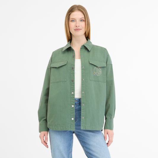 A woman with long hair wears a green button-up LITTA JACKET 2511-60004-5033 from RAGWEAR, featuring two pockets and a white graphic. She combines it with a light green top, blue jeans, and stands against a plain white background.